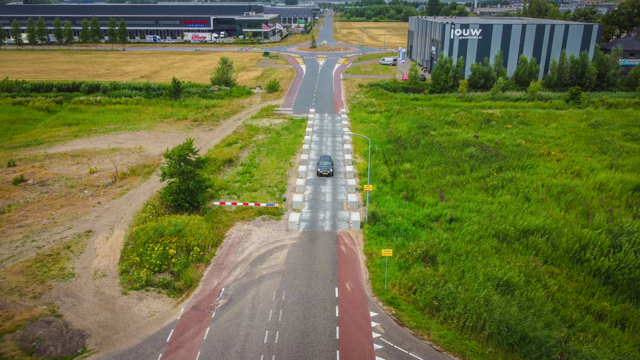 Luchtfoto van auto op smalle weg met betonblokken, richting rotonde bij bedrijfspanden en grasvelden