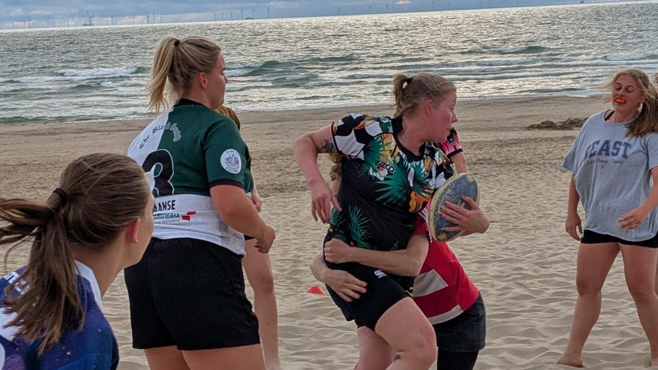 Vrouwen spelen rugby op het strand; een speelster met bal wordt getackeld, met de zee op de achtergrond