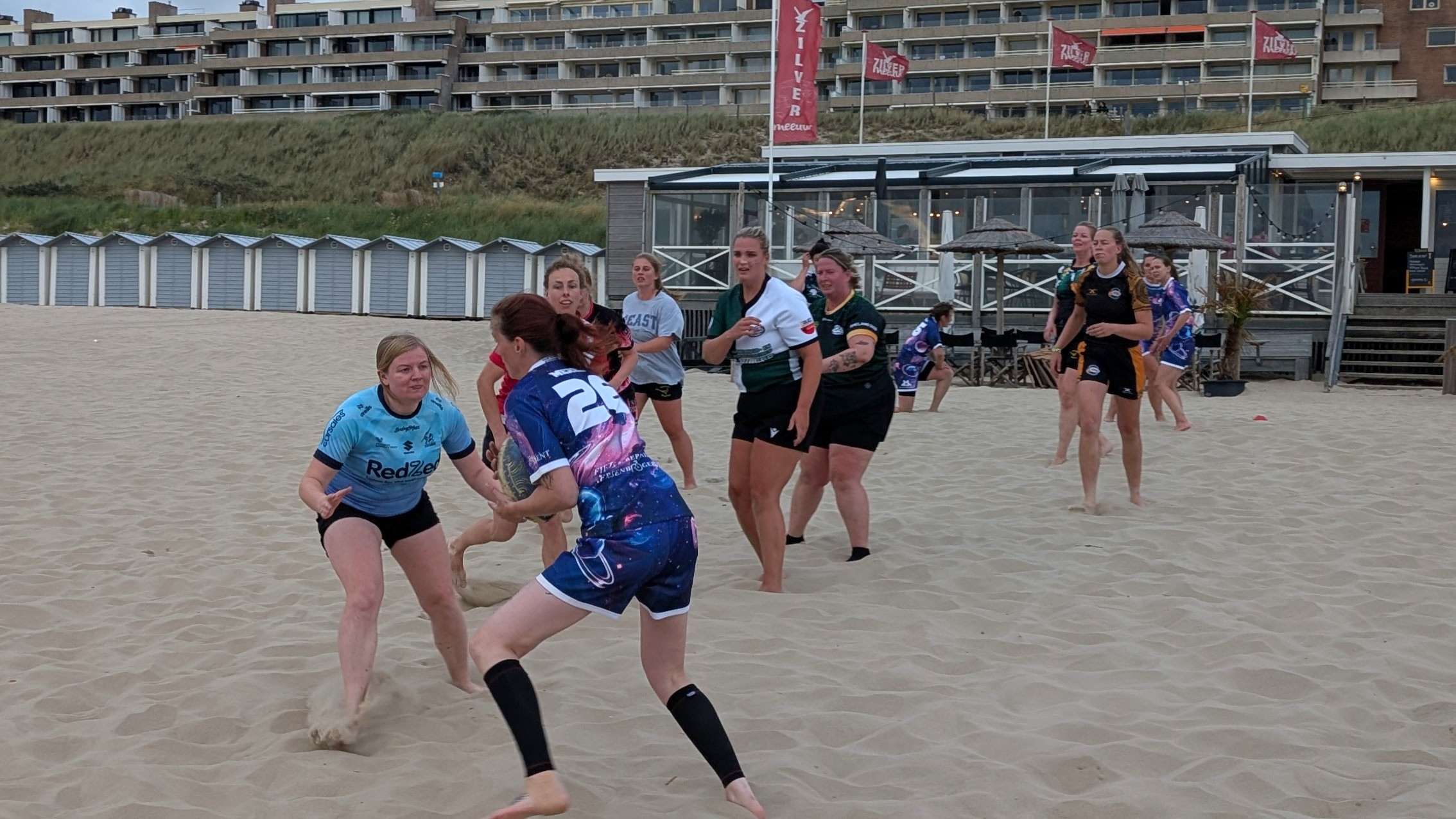 Vrouwen spelen beachrugby op het strand; speelster in paars rent met de bal terwijl een tegenstander in blauw verdedigt