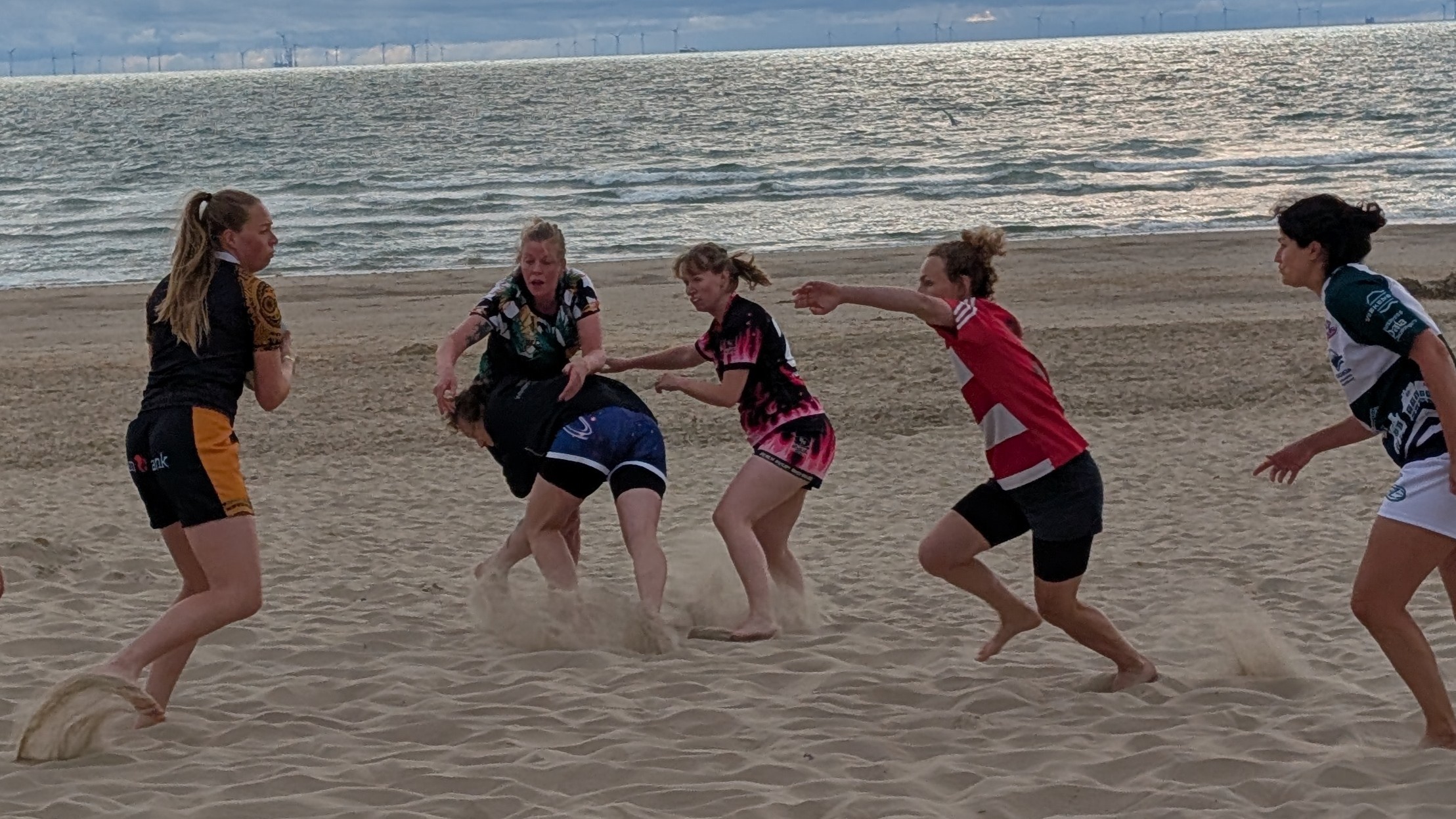Vrouwen spelen rugby op het strand, met een tackle in het zand terwijl teamgenoten toekijken; zee op de achtergrond