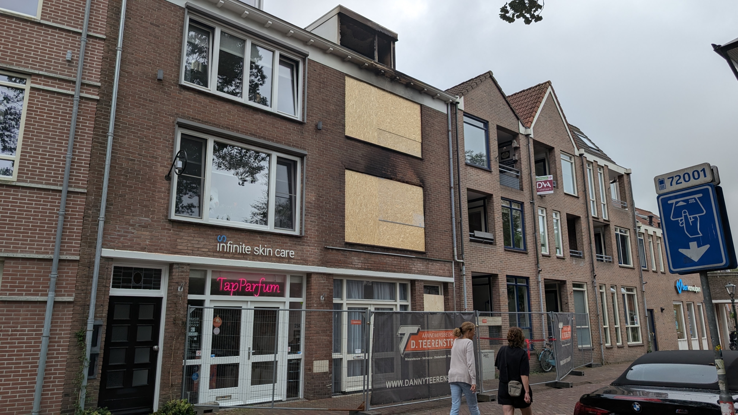 Bakstenen gebouw met dichtgetimmerde ramen en brandschade bij de bovenste verdieping, met hekwerk en twee voorbijgangers