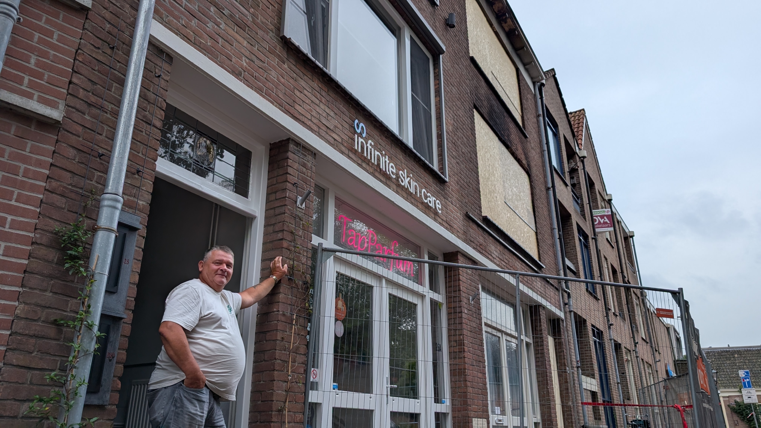 Man staat bij voordeur van bakstenen pand met gevelbordje Infinite skin care en raamtekst TapParfum, achter bouwhek