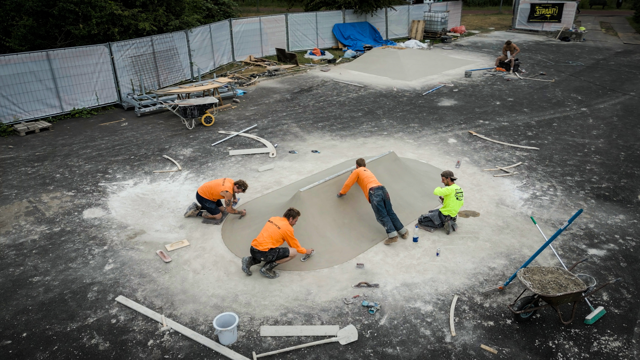 Bouwvakkers smeren beton glad in een skateparkbowl, met gereedschap en kruiwagen op een afgezette bouwplaats