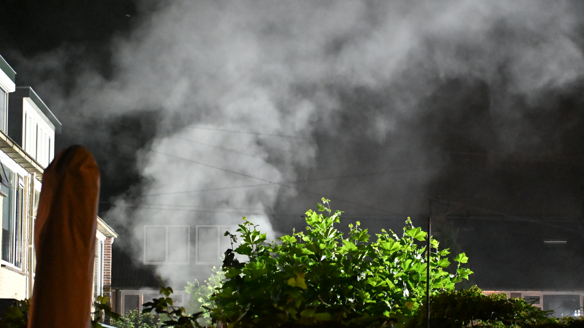 Dikke rookwolken stijgen ’s nachts op achter huizen, met fel verlichte groene struiken op de voorgrond