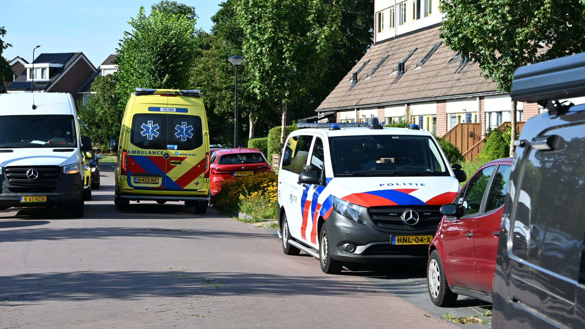 Politieauto en ambulance geparkeerd in een woonstraat met huizen en bomen, met meerdere auto’s langs de weg