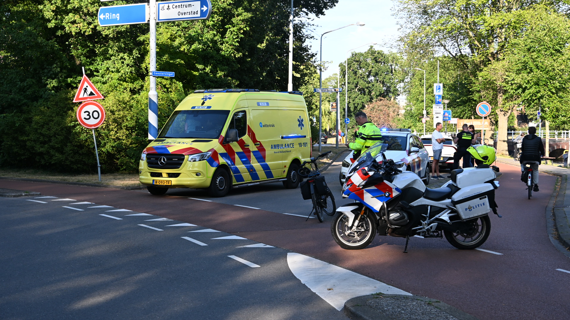 Ambulance en politiemotor blokkeren een kruispunt; agenten staan bij een politieauto en wegwijzers naar Ring en Centrum