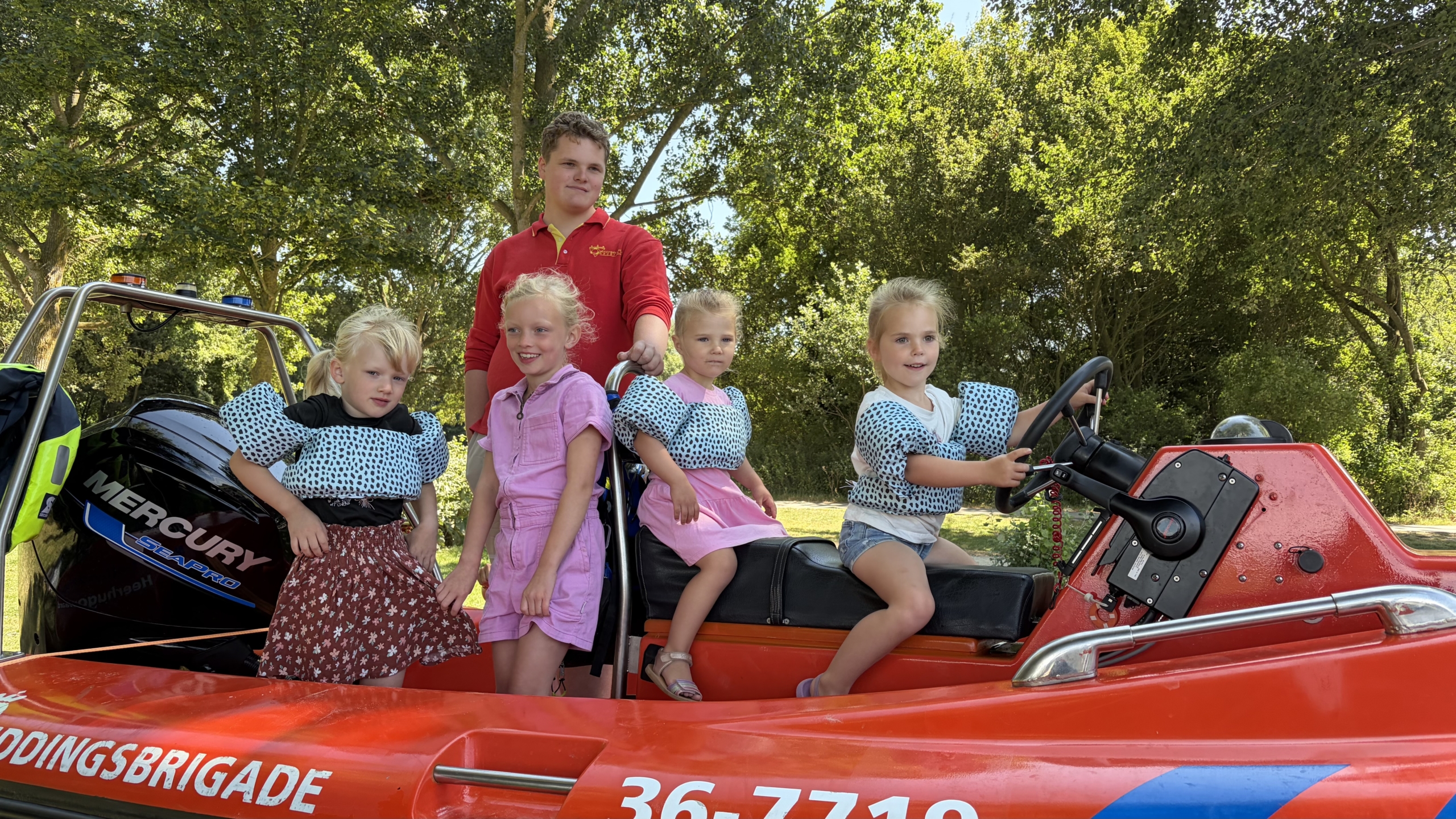 Vier kinderen met zwemvesten op een reddingsboot, met een tiener in rood erachter, in een groene omgeving