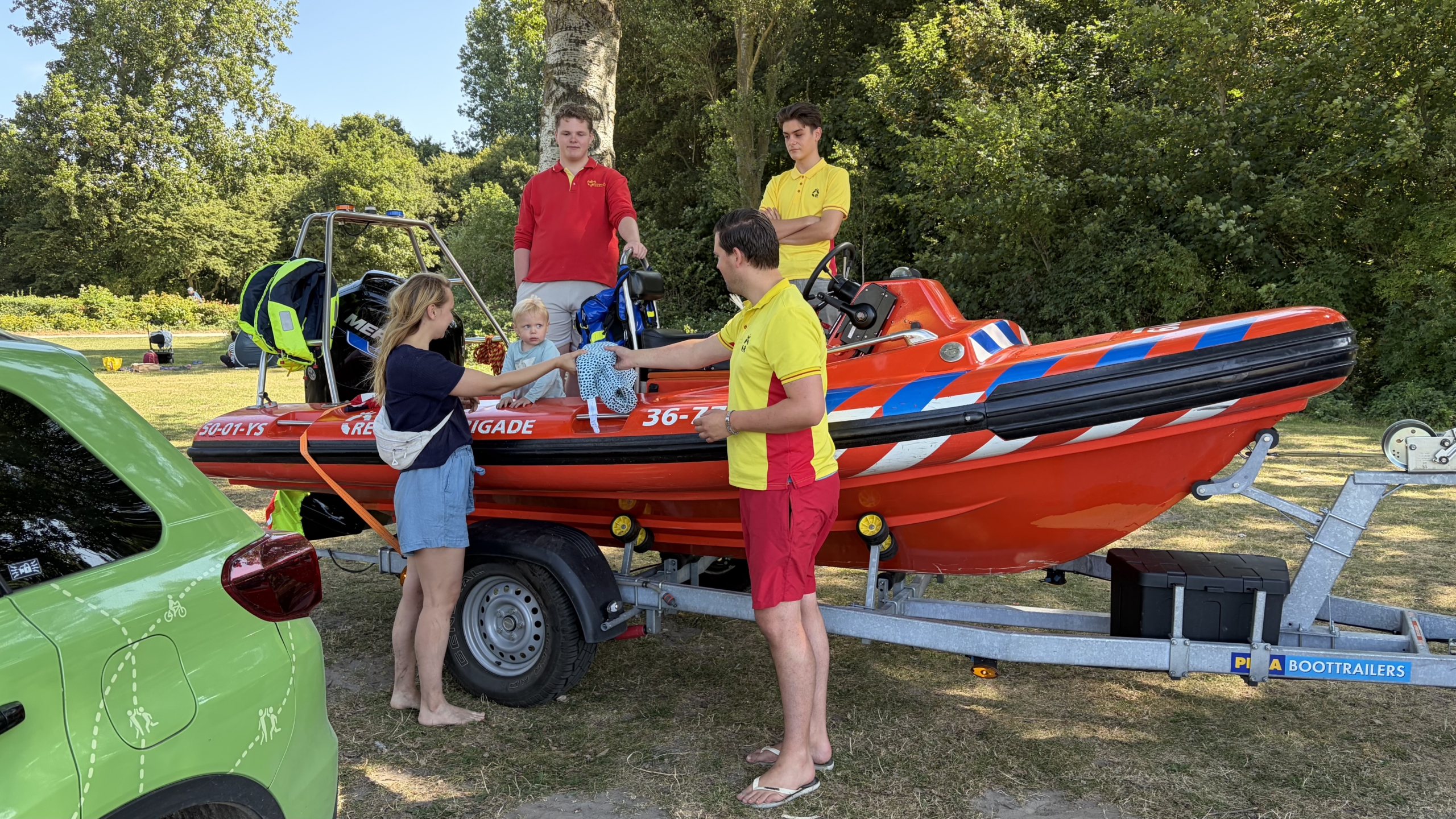 Twee reddingsbrigadeleden bij een rood rescuebootje op trailer geven een drijfvest aan een vrouw met kind in de boot