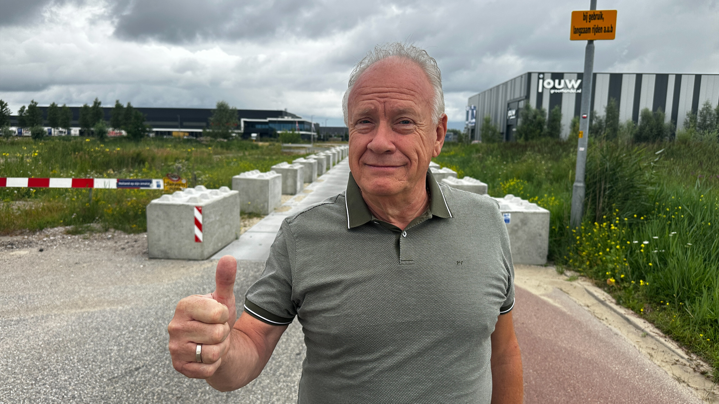Man geeft duim omhoog bij wegafzetting met betonnen blokken, met industriegebouwen en bewolkte lucht op achtergrond