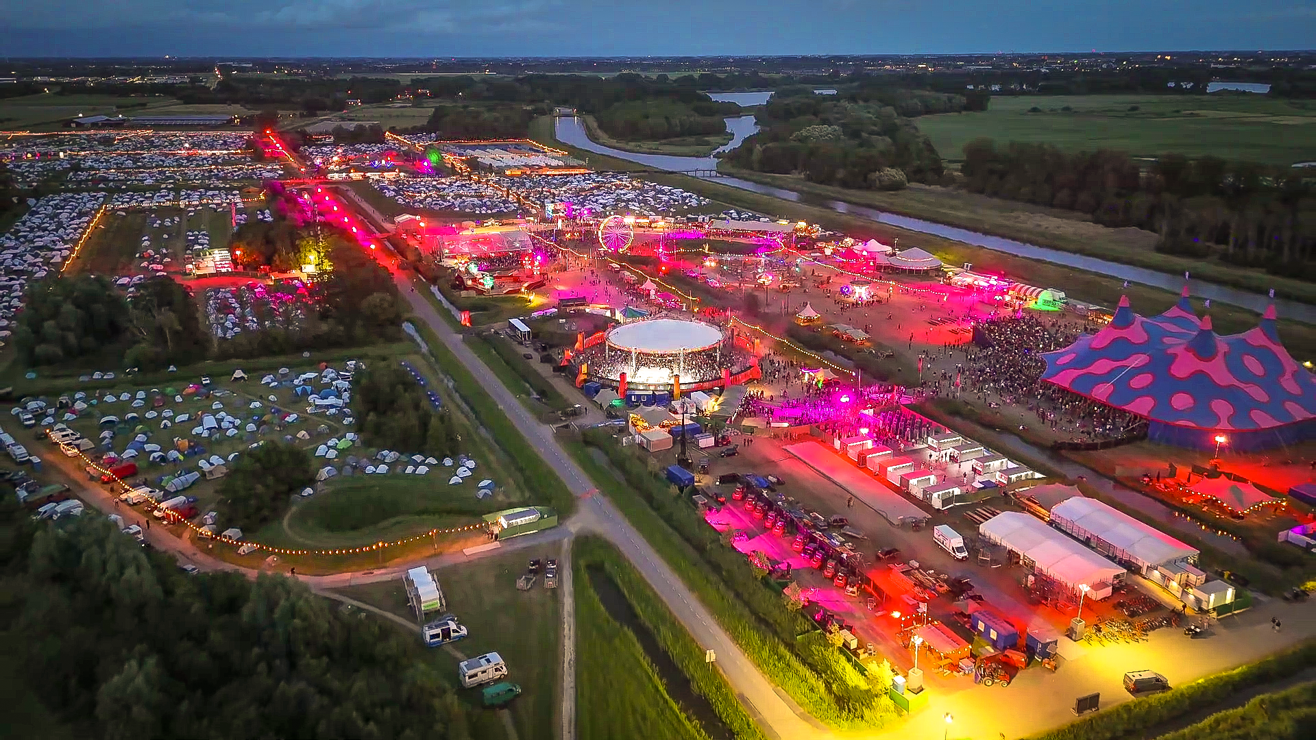 Luchtfoto van een verlicht festivalterrein bij schemering met tentencamping, mensenmassa en rivier in het landschap