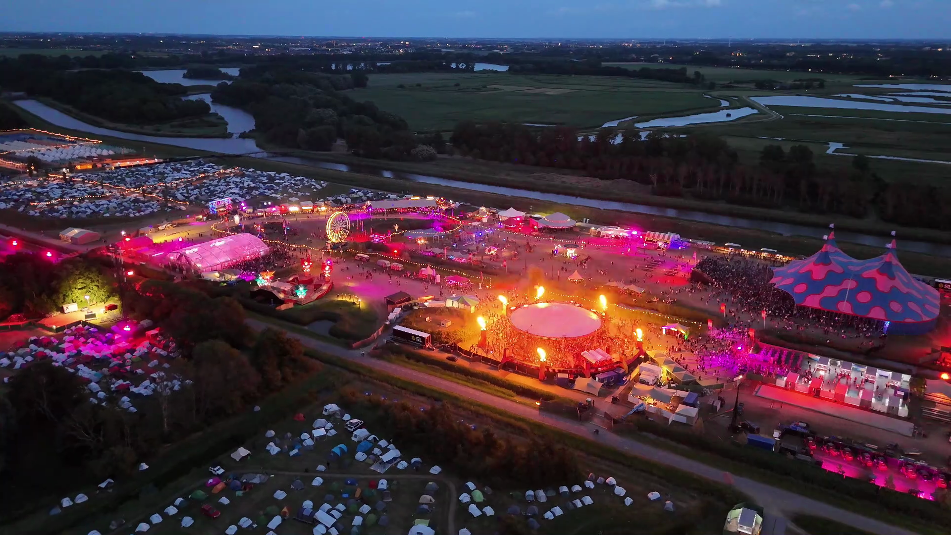 Luchtfoto van festivalterrein in de schemering met felgekleurde lichten, groot tentdak, reuzenrad en camping eromheen