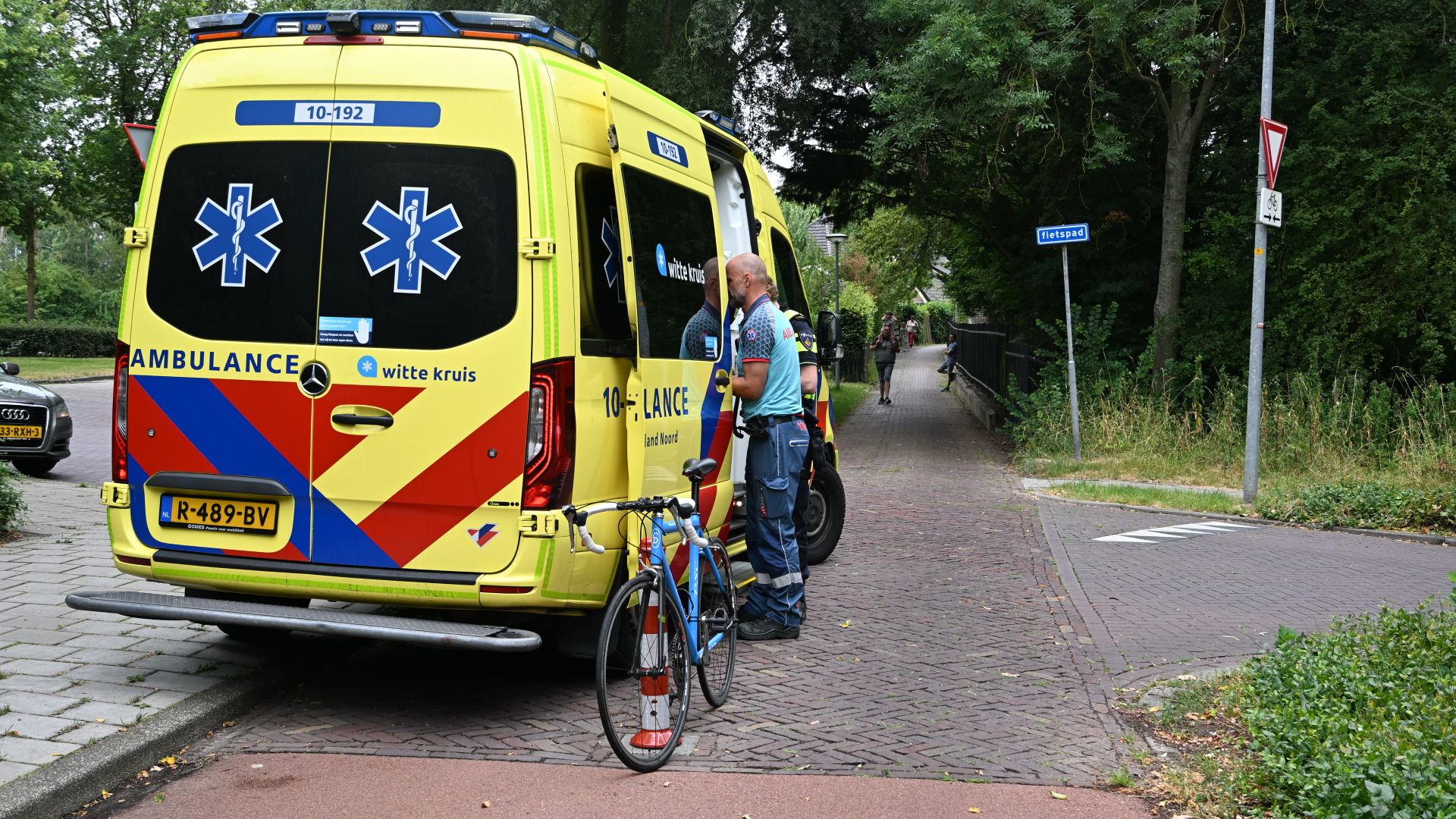 Ambulance van Witte Kruis staat bij fietspad; hulpverlener naast een fiets en afzetkegel, voetgangers op de achtergrond