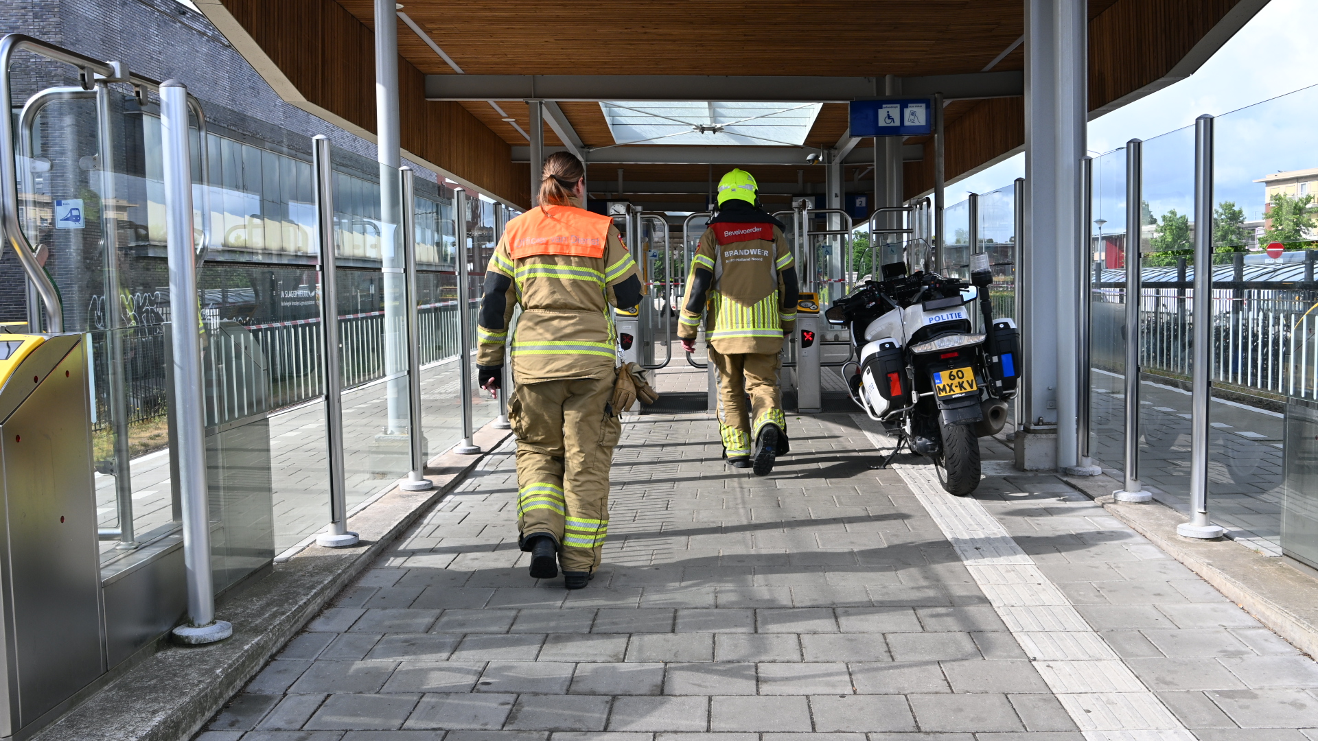 Twee brandweerlieden lopen over een tramhalte richting poortjes; rechts staat een politiemotor geparkeerd