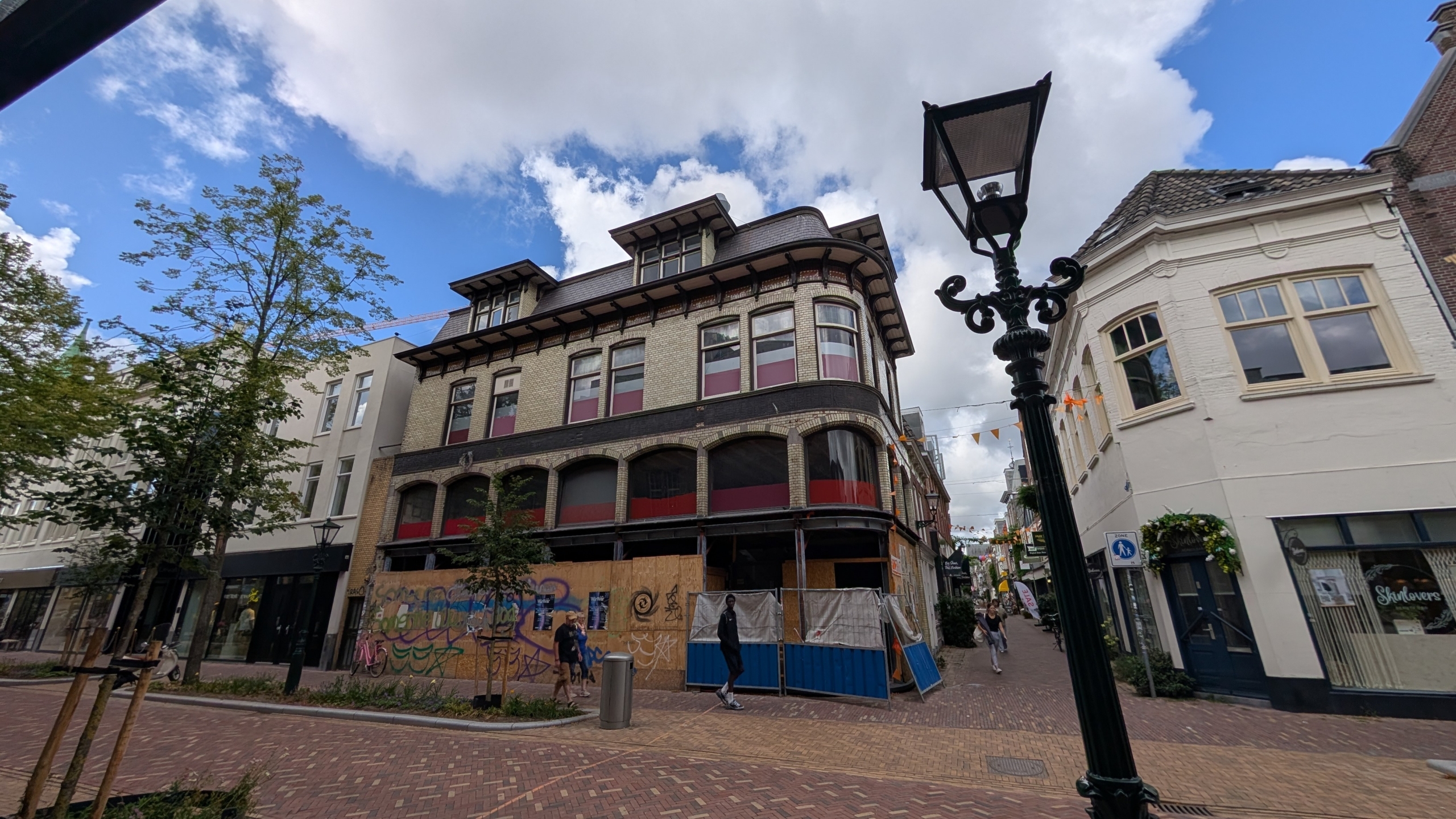 Straatbeeld met historisch pand in renovatie, afgeschermd met houten platen en bouwhekken, onder blauwe lucht met lantaarnpaal