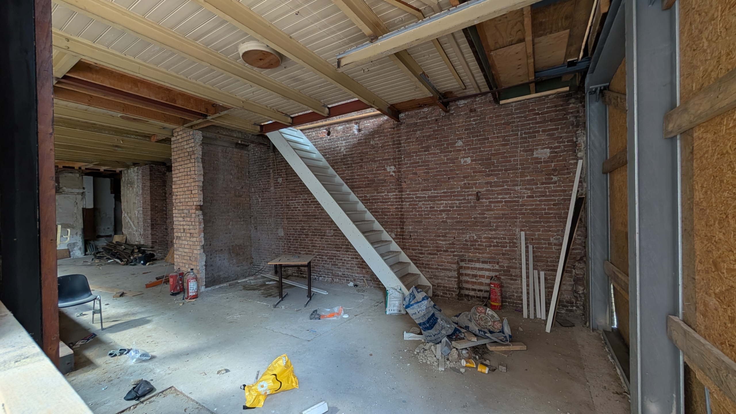Lege ruimte in verbouwing met bakstenen muur, metalen plafondbalken en witte trap, met rommel op de vloer