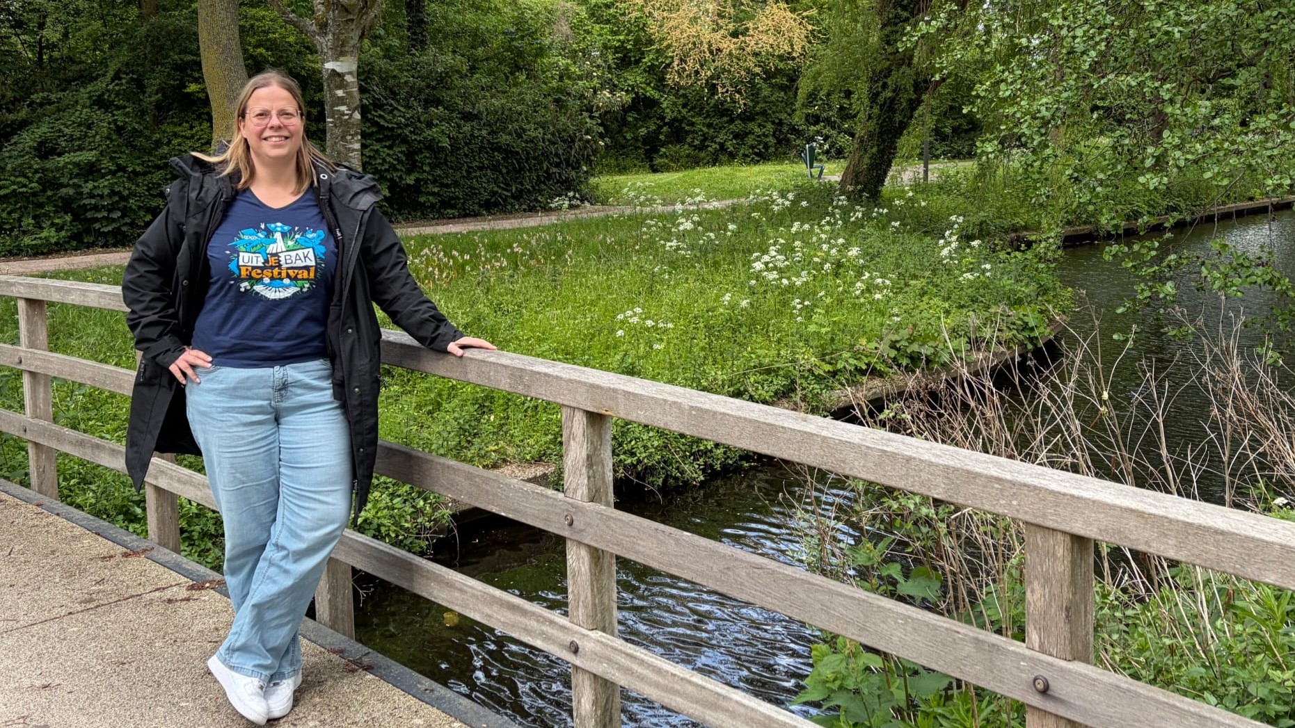 Vrouw met bril leunt op houten brugleuning bij een sloot in groen park, met T-shirttekst “Uit je bak Festival”