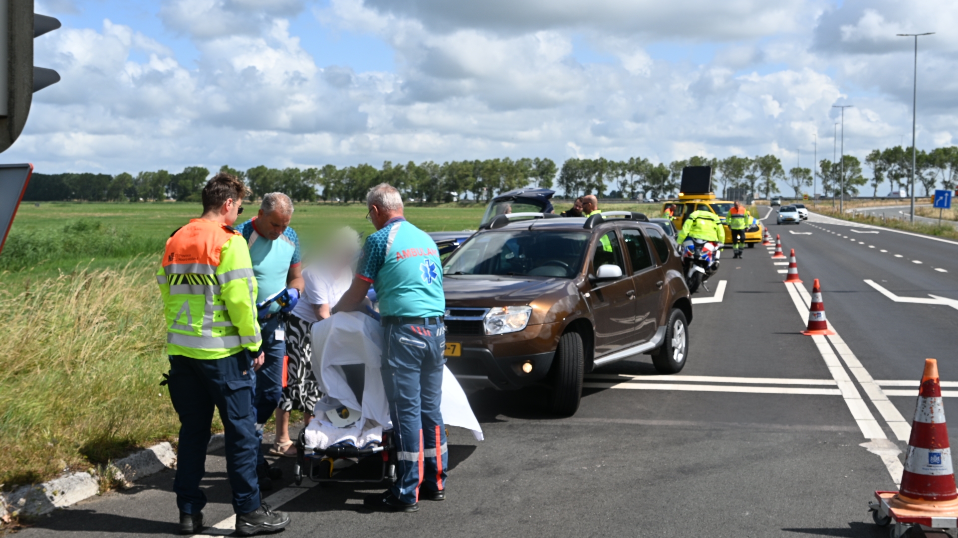 Hulpverleners verzorgen een slachtoffer naast een auto op een afgezet wegvak met pionnen en hulpdiensten op de achtergrond