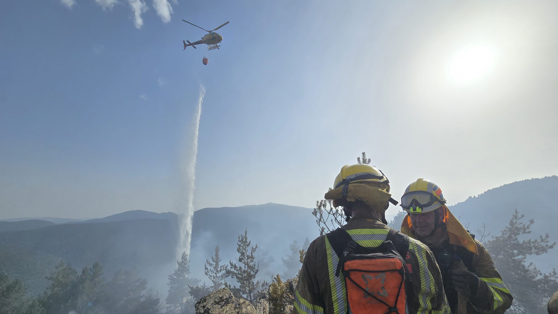 Twee brandweerlieden kijken toe terwijl een helikopter met waterzak water dropt boven een rokerig bosgebied