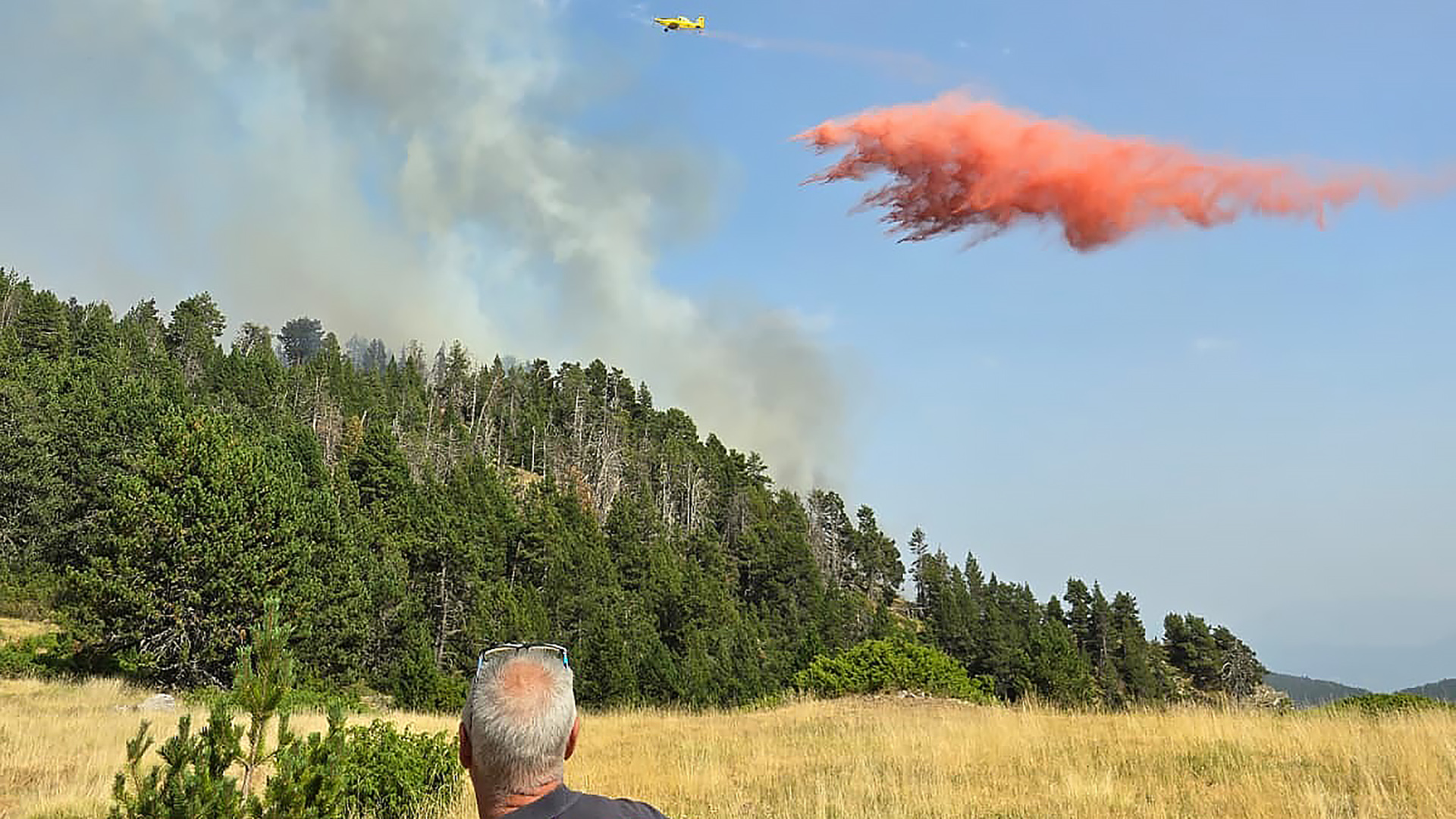 Klein vliegtuig dropt rood brandvertrager boven een bosbrand; rook boven heuvels, man kijkt toe vanaf veld