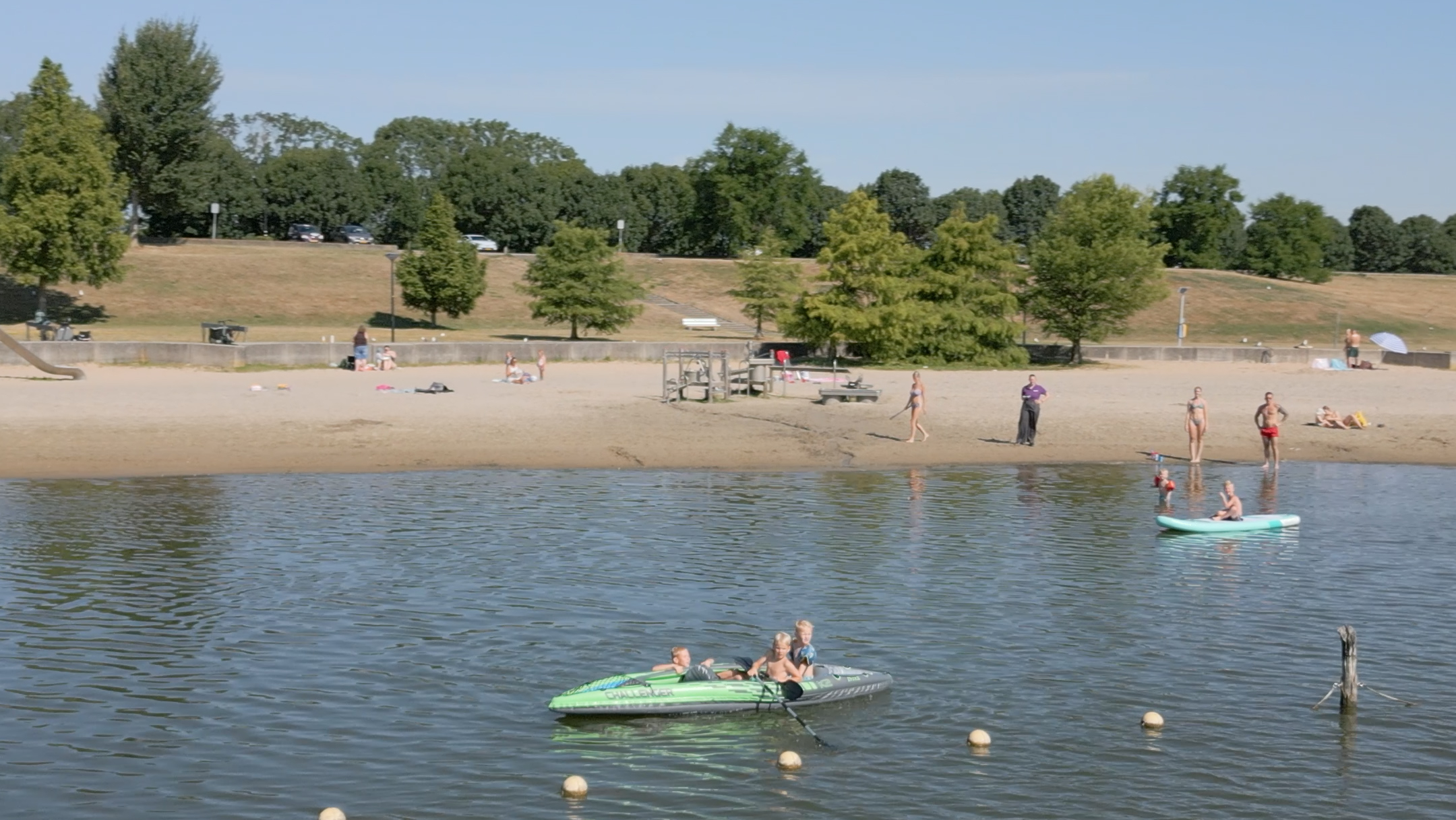 Kinderen in een opblaasboot op een meer, met een zandstrand en badgasten op de achtergrond
