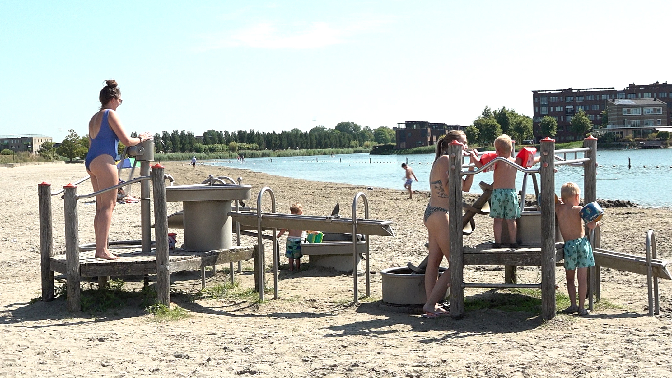 Mensen in zwemkleding gebruiken buitendouches op een zandstrand bij een recreatieplas, met gebouwen op de achtergrond