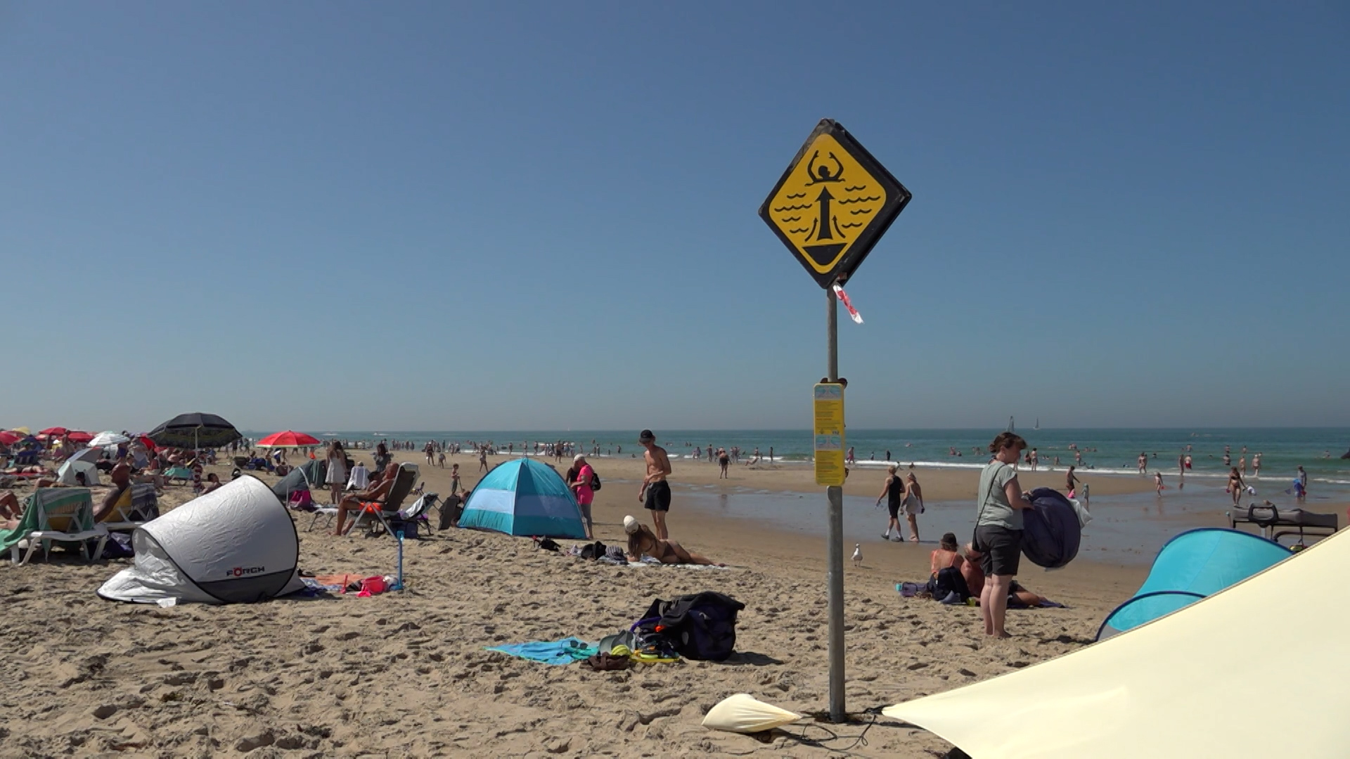 Druk strand met zwemmers en parasols, met op de voorgrond een geel waarschuwingsbord voor stroming in zee