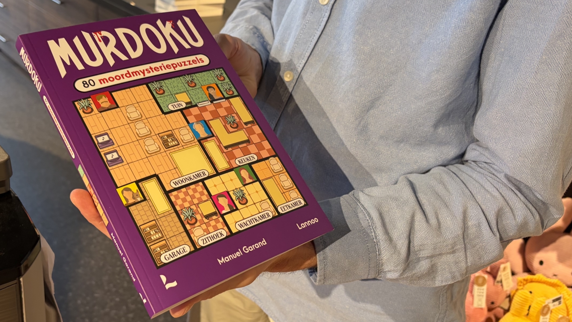 Persoon houdt het paarse puzzelboek Murdoku vast, met de tekst 80 moordmysteriepuzzels en een plattegrond op de cover