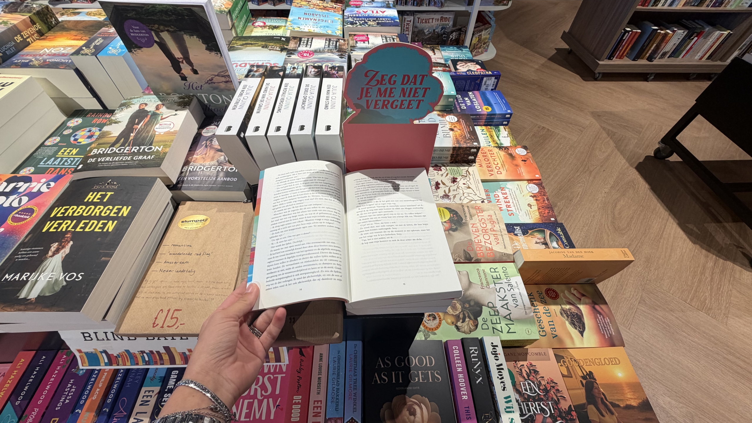 Hand houdt open boek boven tafel vol romans in boekwinkel, met bordje Zeg dat je me niet vergeet