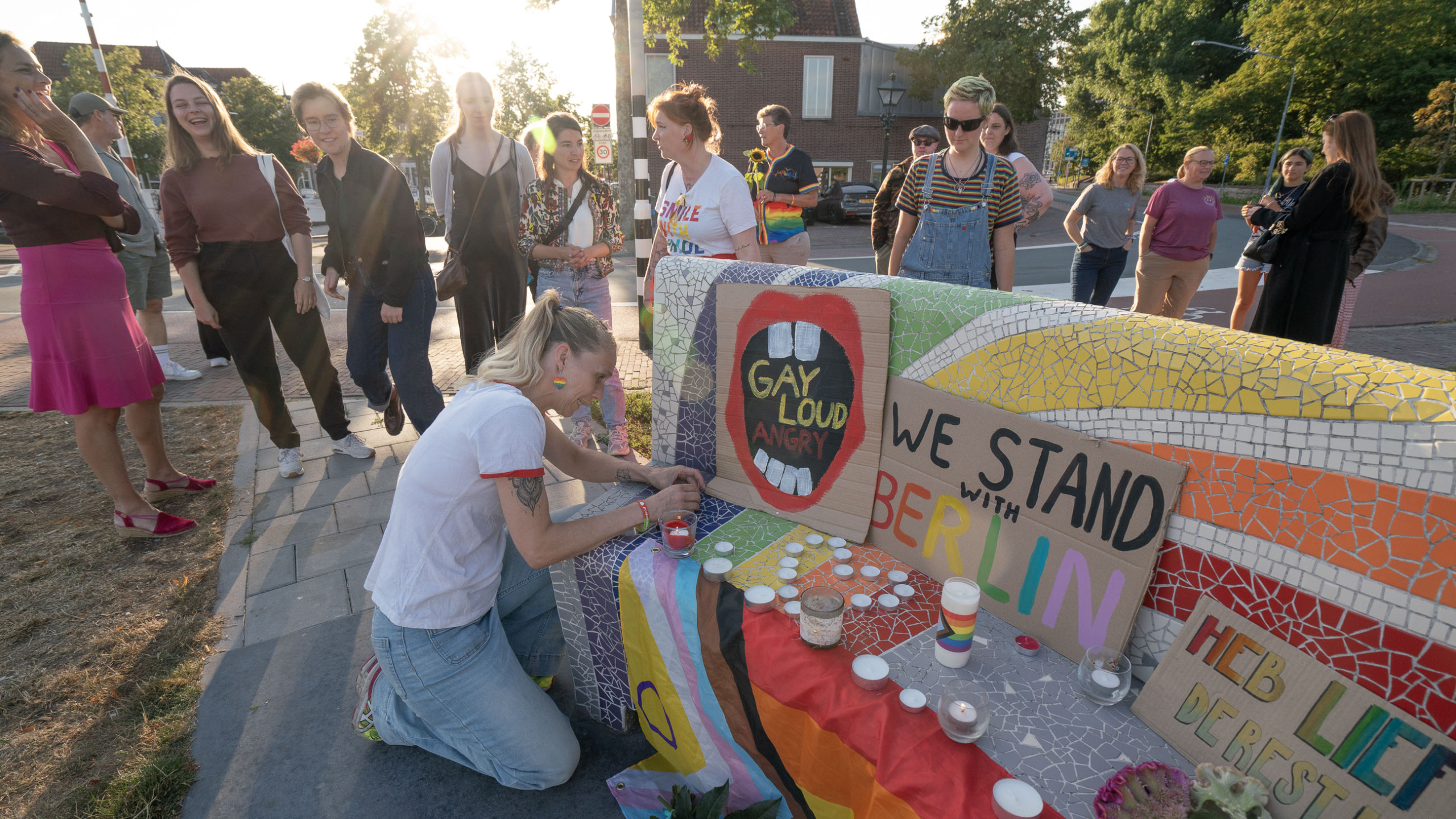 Groep mensen bij een regenboogbank met kaarsen en borden met teksten als We stand with Berlin en Gay Loud Angry