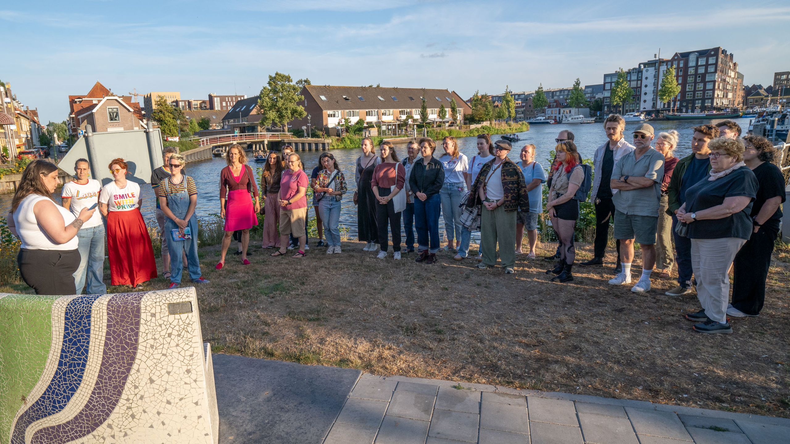 Groep mensen luistert bij een kanaal in de stad, met boten en woonhuizen op de achtergrond