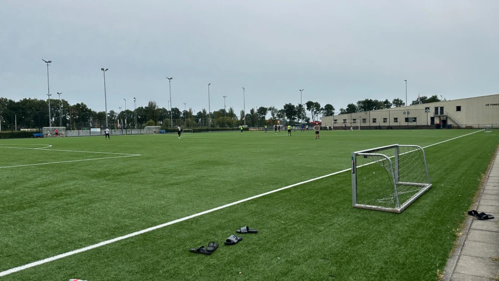 Voetbalveld met spelers op de achtergrond, een klein doel en slippers op de voorgrond naast de zijlijn