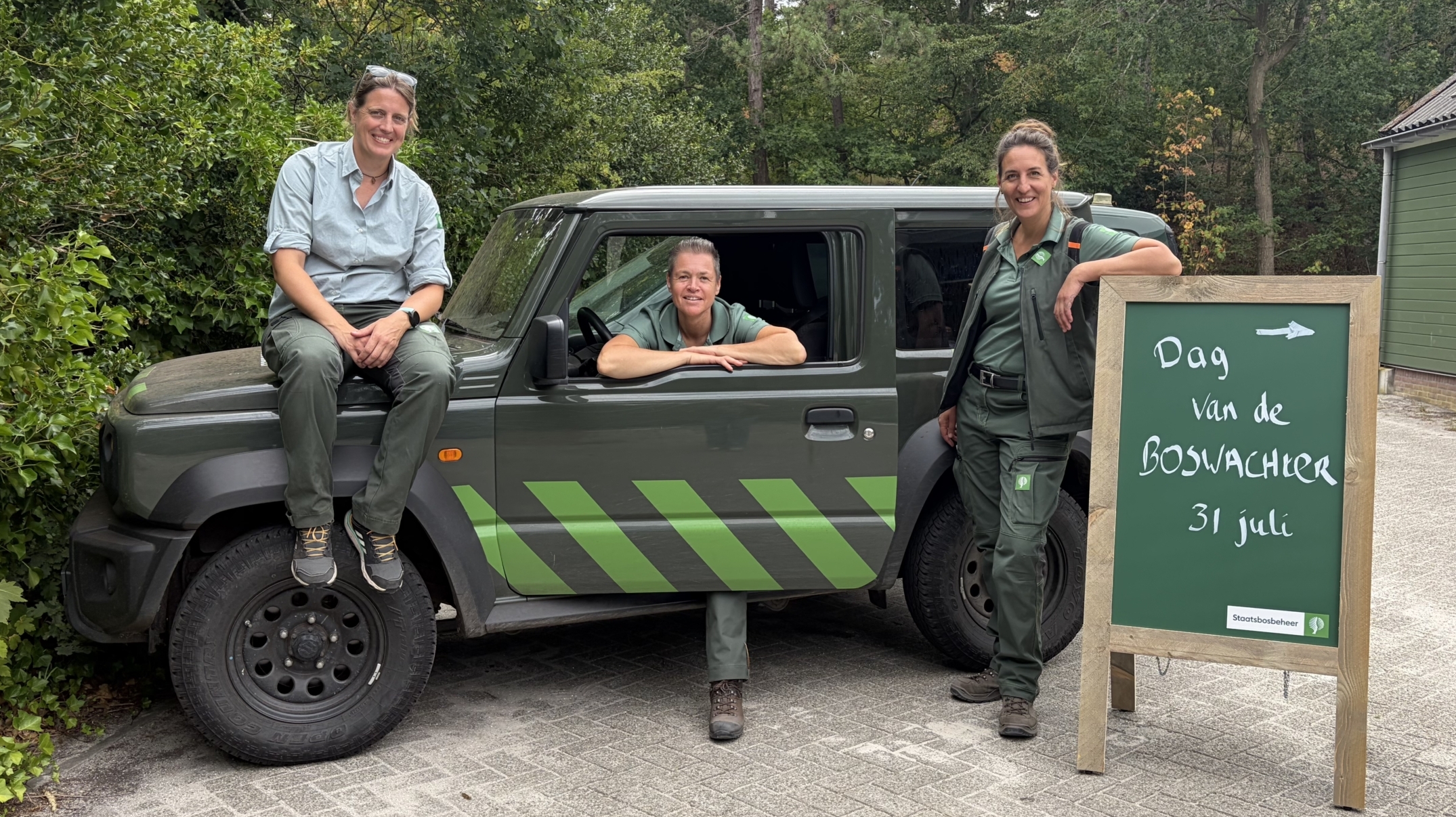 Drie boswachters poseren bij een groen dienstvoertuig naast een bord met tekst “Dag van de boswachter 31 juli”