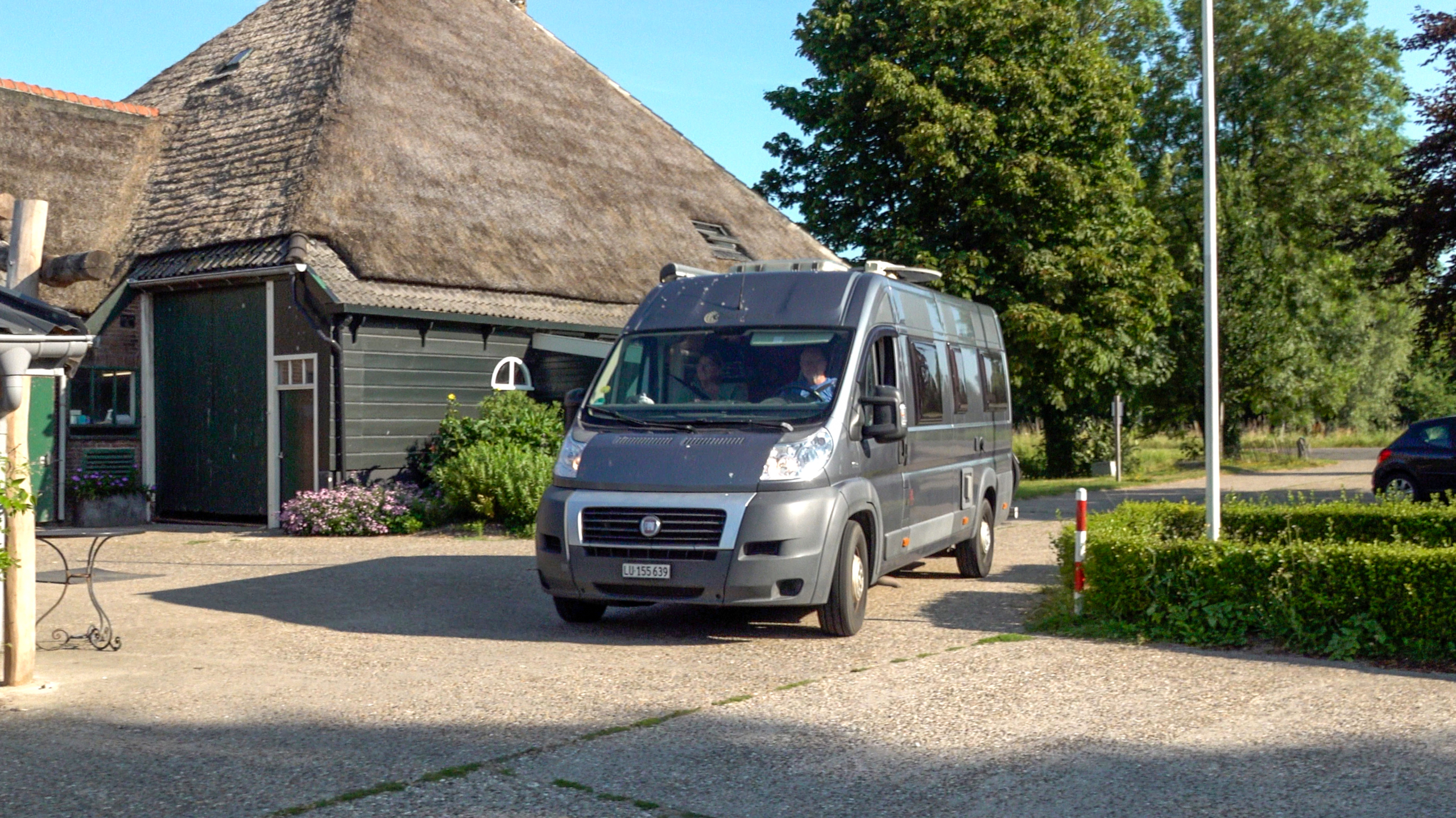 Grijze camperbus geparkeerd voor een boerderij met rieten dak, met bomen en groen op de achtergrond
