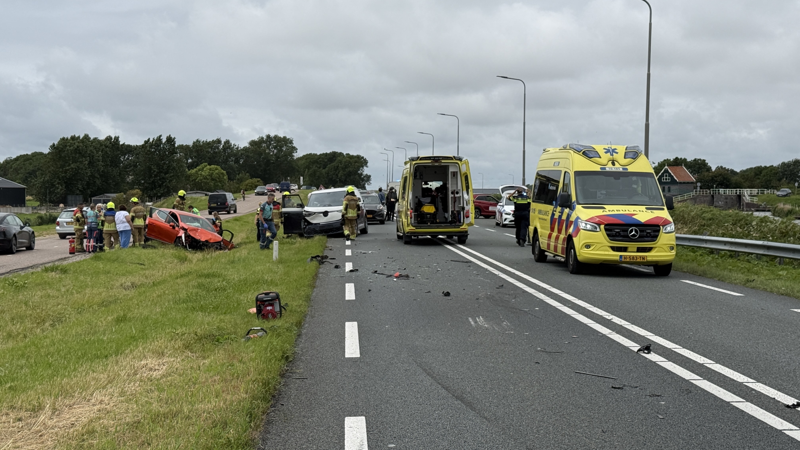 Verkeersongeval op een snelweg met betrokken auto's, brandweerlieden en twee ambulances aanwezig, omstanders kijken toe.