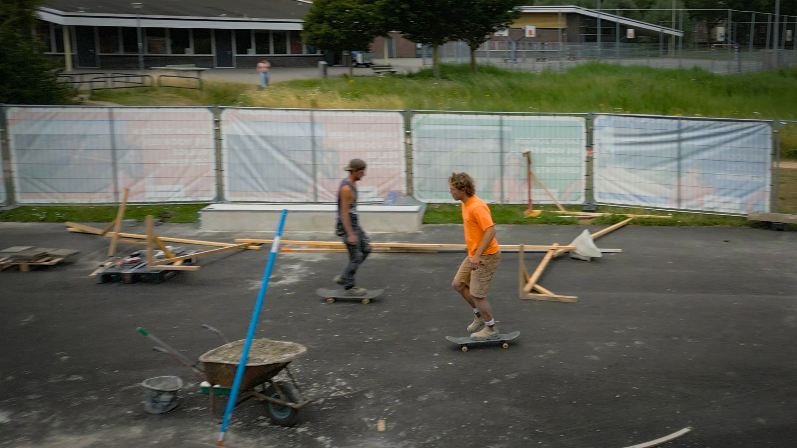 Twee skaters rijden over een asfaltterrein met verspreid hout en een kruiwagen, achter een bouwhek