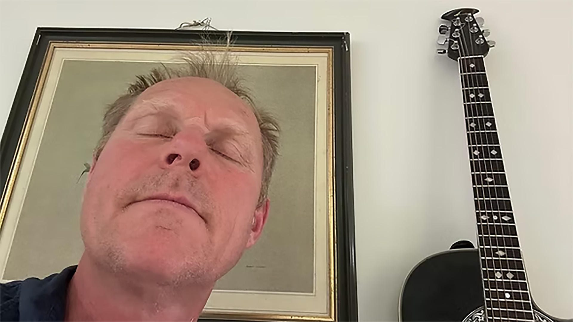 Close-up van een man met gesloten ogen, met een ingelijste afbeelding achter hem en een gitaar rechts in beeld