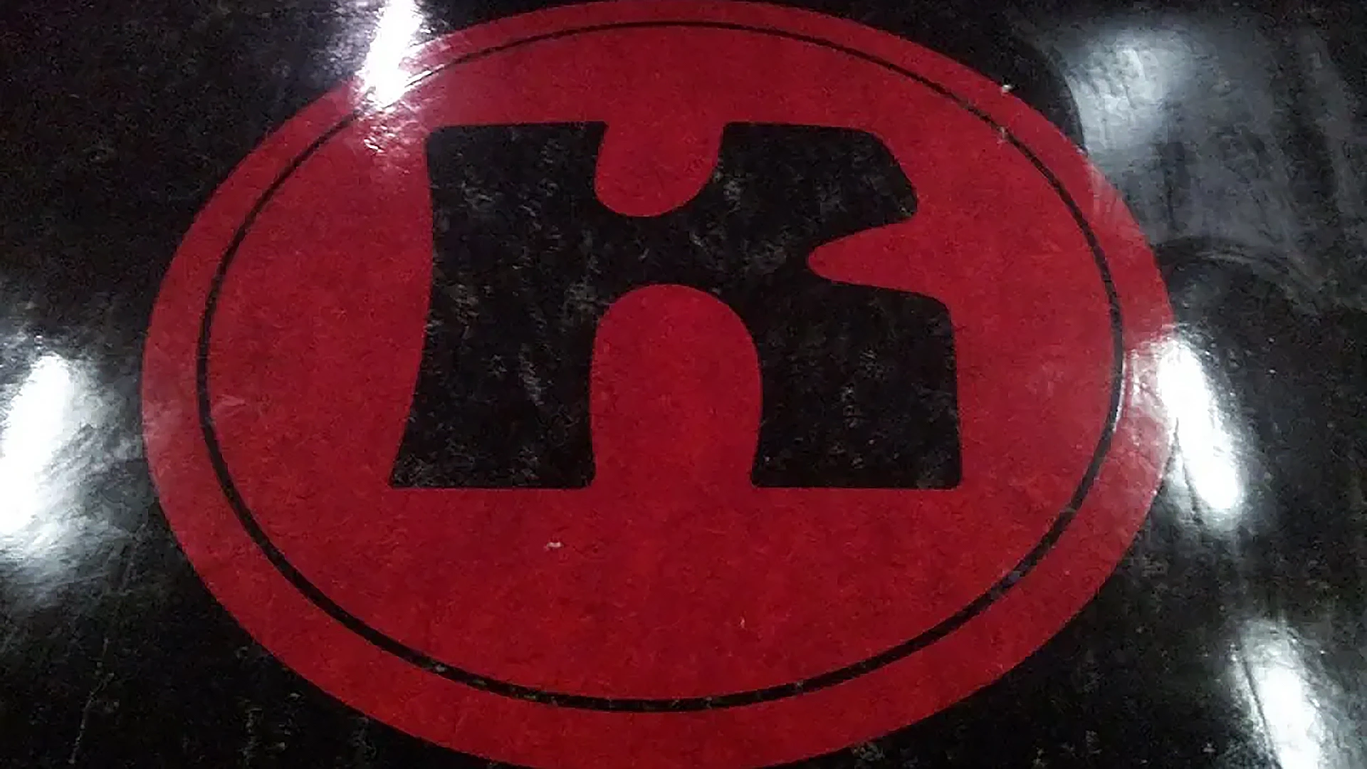 Rood ovaal logo met zwarte, gestileerde hoofdletter K op een glanzende zwarte ondergrond