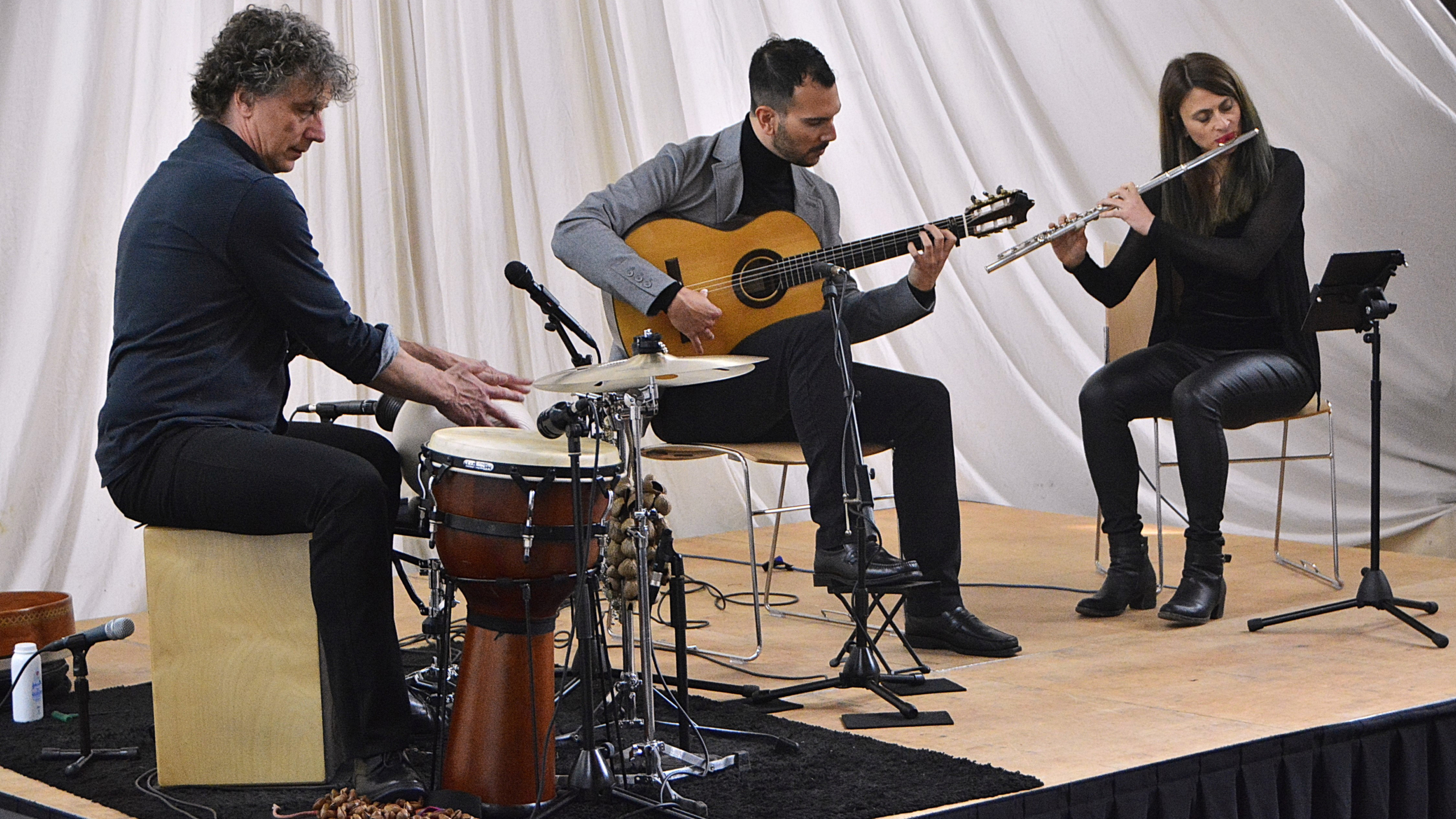 Drie muzikanten op een podium: percussionist op cajón, gitarist en fluitiste met lessenaar en microfoons