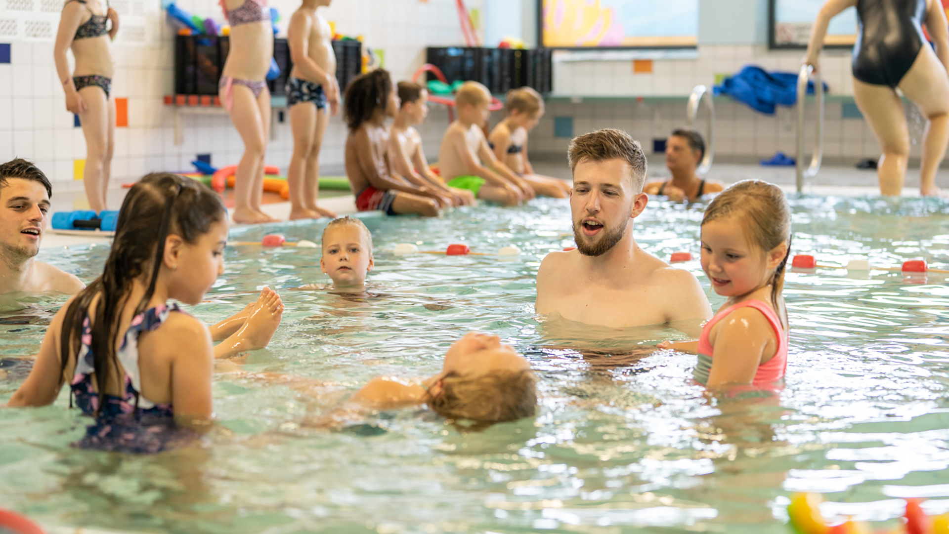 Kinderen en een instructeur in een zwembad zijn bezig met zwemlessen, omringd door andere kinderen die toekijken.