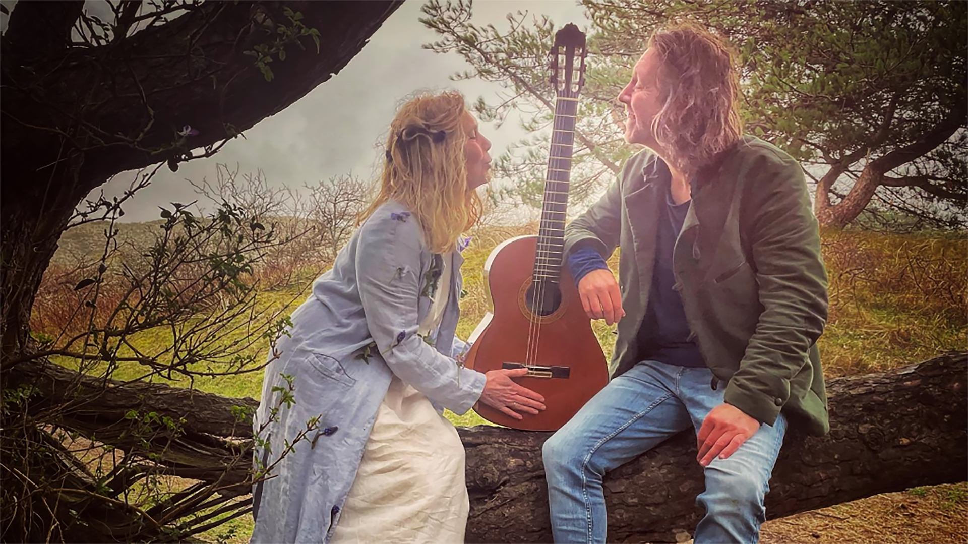 Man en vrouw zitten op een boomstam in de natuur, naast een klassieke gitaar, kijkend naar elkaar met een glimlach.