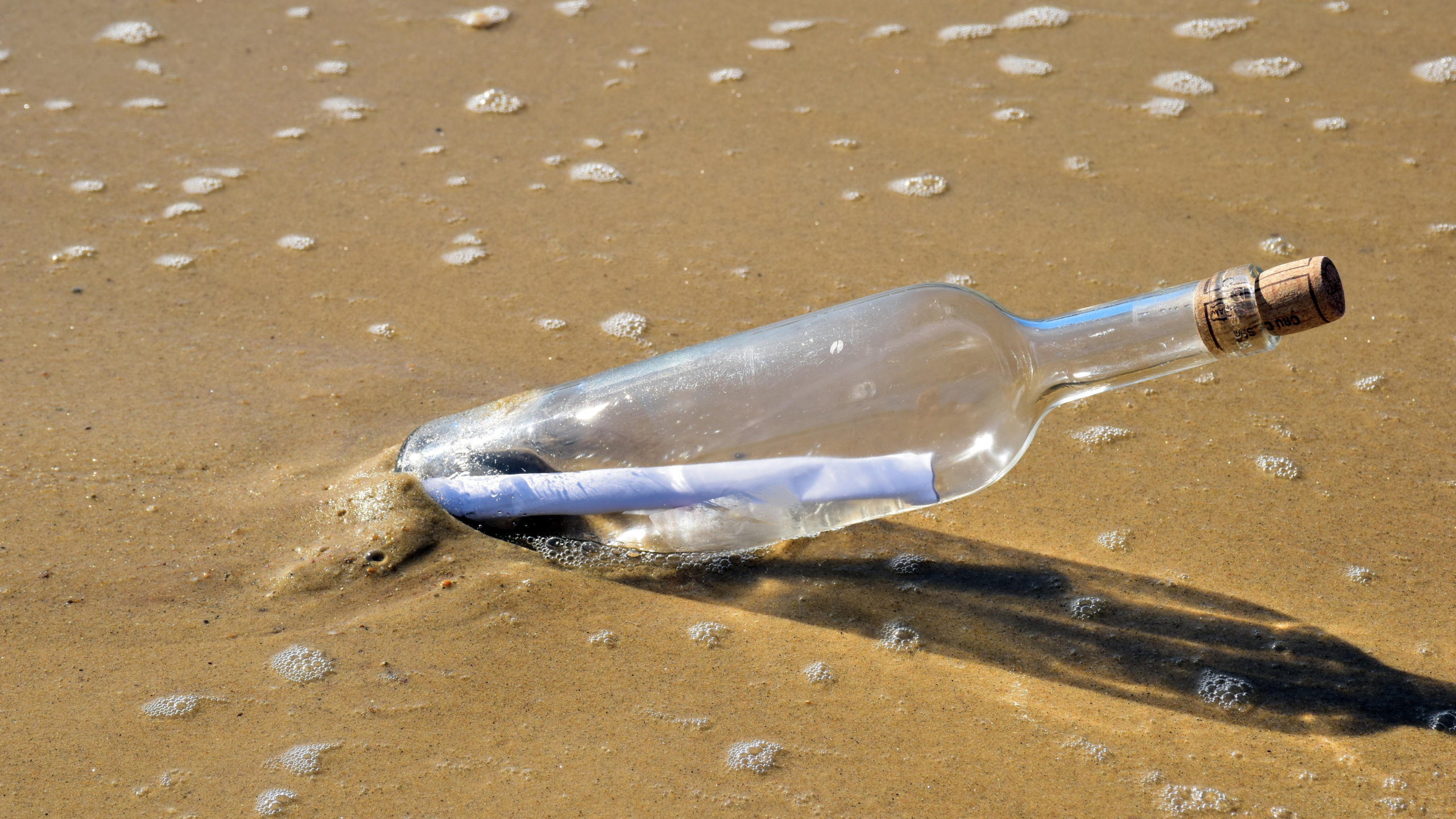 Glazen fles met kurk en briefje erin ligt aangespoeld op nat zand met wat zeeschuim