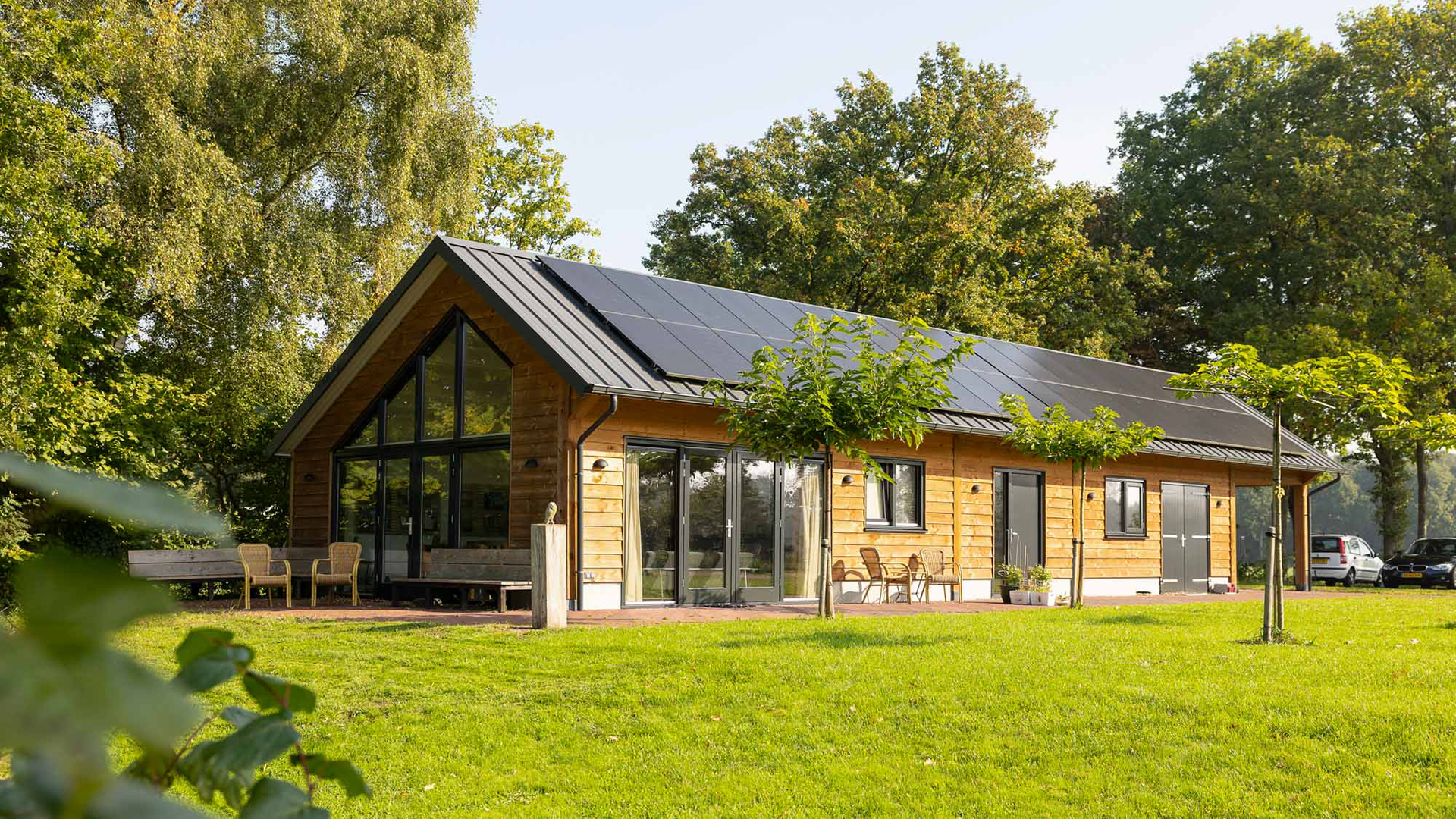 Houten vakantiewoning met grote glazen pui en zonnepanelen op het dak, aan een groen grasveld met bomen
