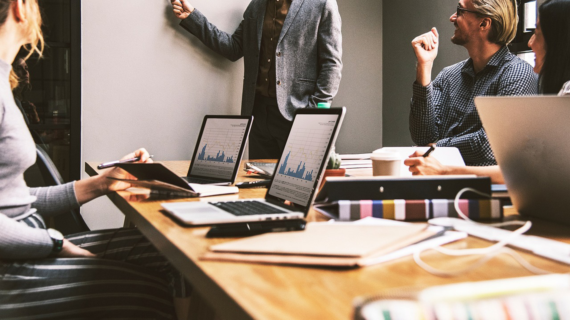 Collega’s in overleg rond een vergadertafel met laptops waarop grafieken te zien zijn, terwijl iemand aan een whiteboard presenteert