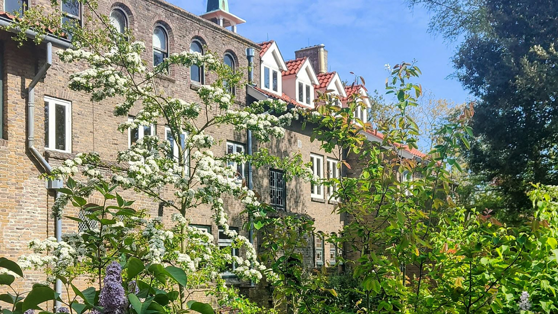 Bakstenen gebouw met rode daken, omlijst door bloeiende bomen en groen, onder een heldere blauwe lucht.