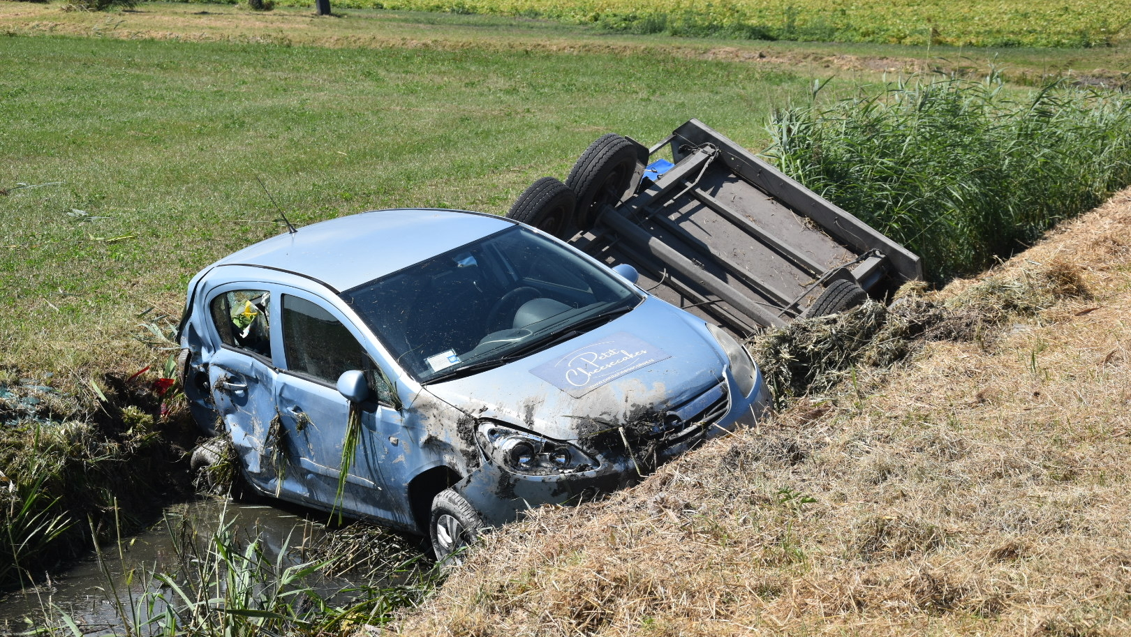 Beschadigde auto in een sloot naast een omgekantelde aanhanger in een landelijk gebied