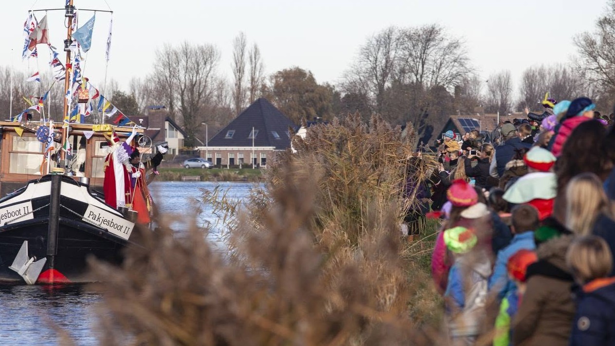 Sinterklaasboot vaart langs oever met riet, terwijl een menigte toeschouwers in winterkleding toekijkt