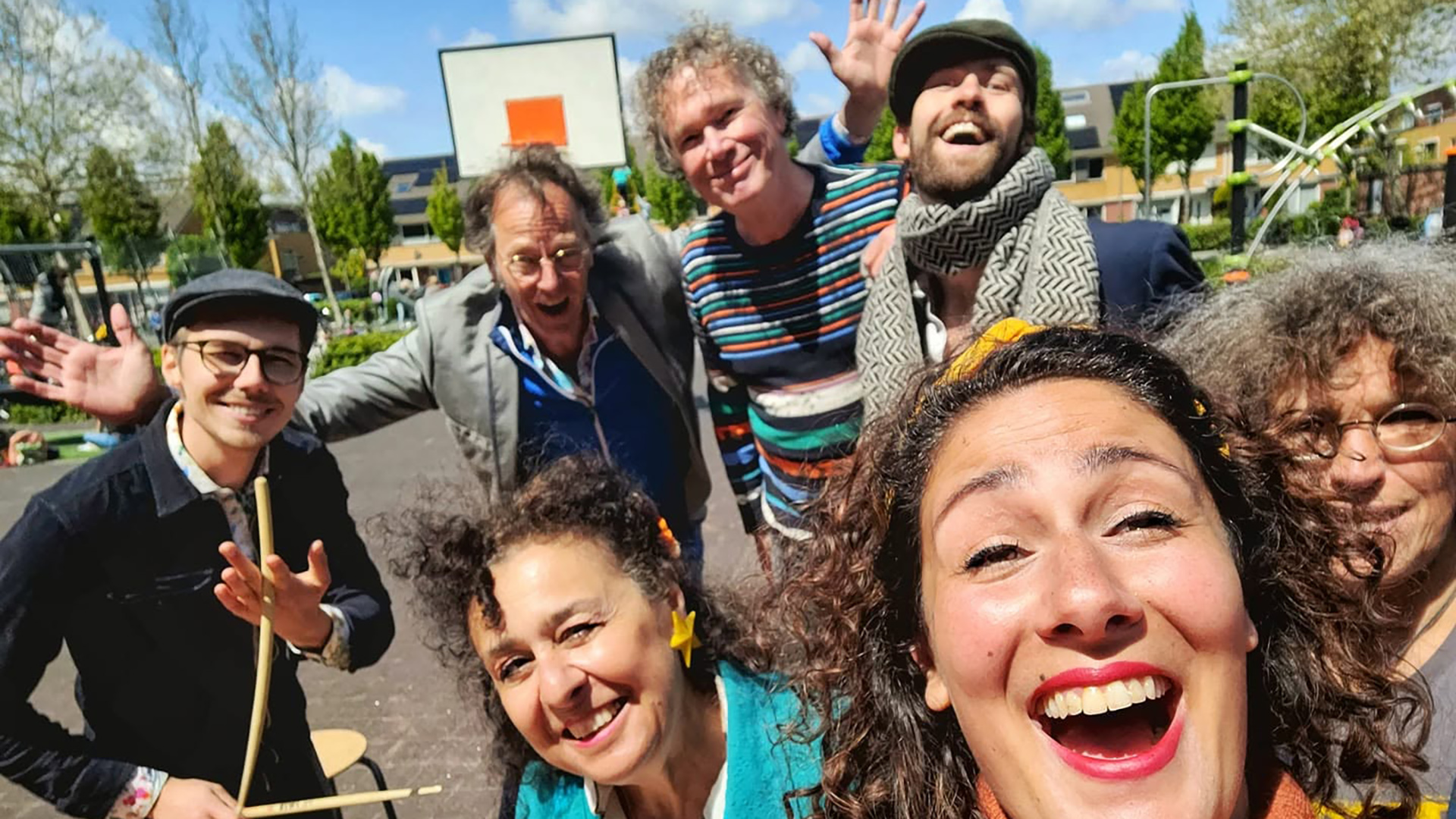 Groepsselfie van lachende vrienden op een speelplein bij een basketbalbord, met iemand die drumstokken vasthoudt