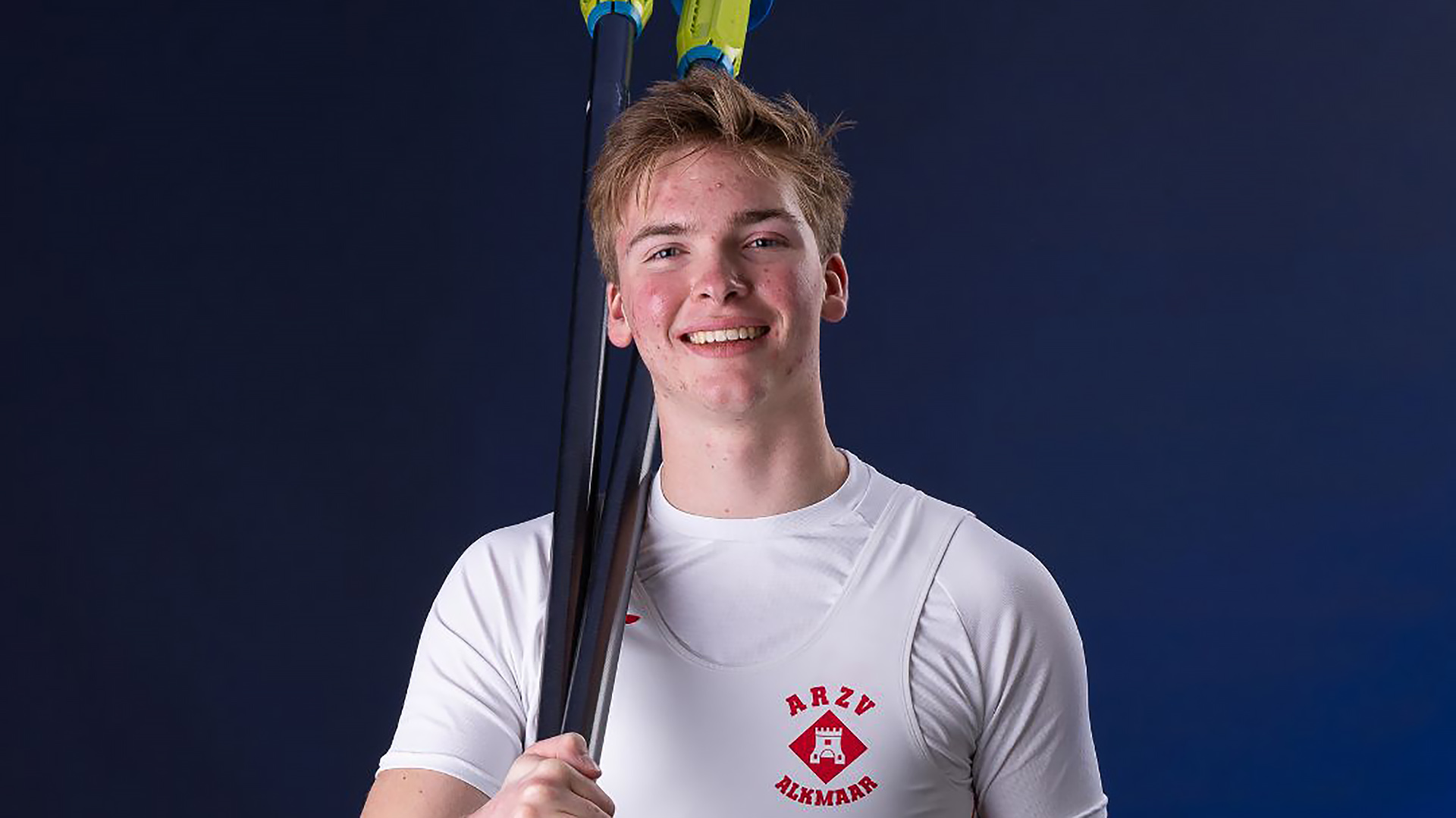 Lachende roeier in wit shirt met ARZV Alkmaar-logo, met roeiriem over de schouder tegen donkerblauwe achtergrond