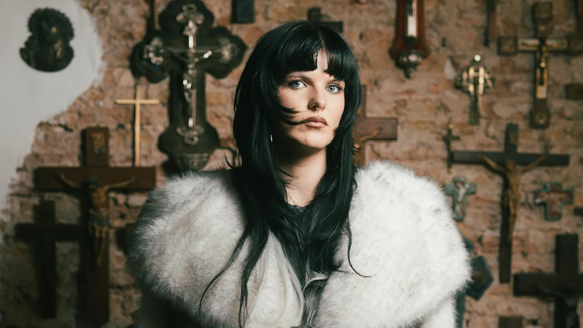 Portret van vrouw met zwart haar en witte bontjas, voor een muur met meerdere kruisen en religieuze ornamenten