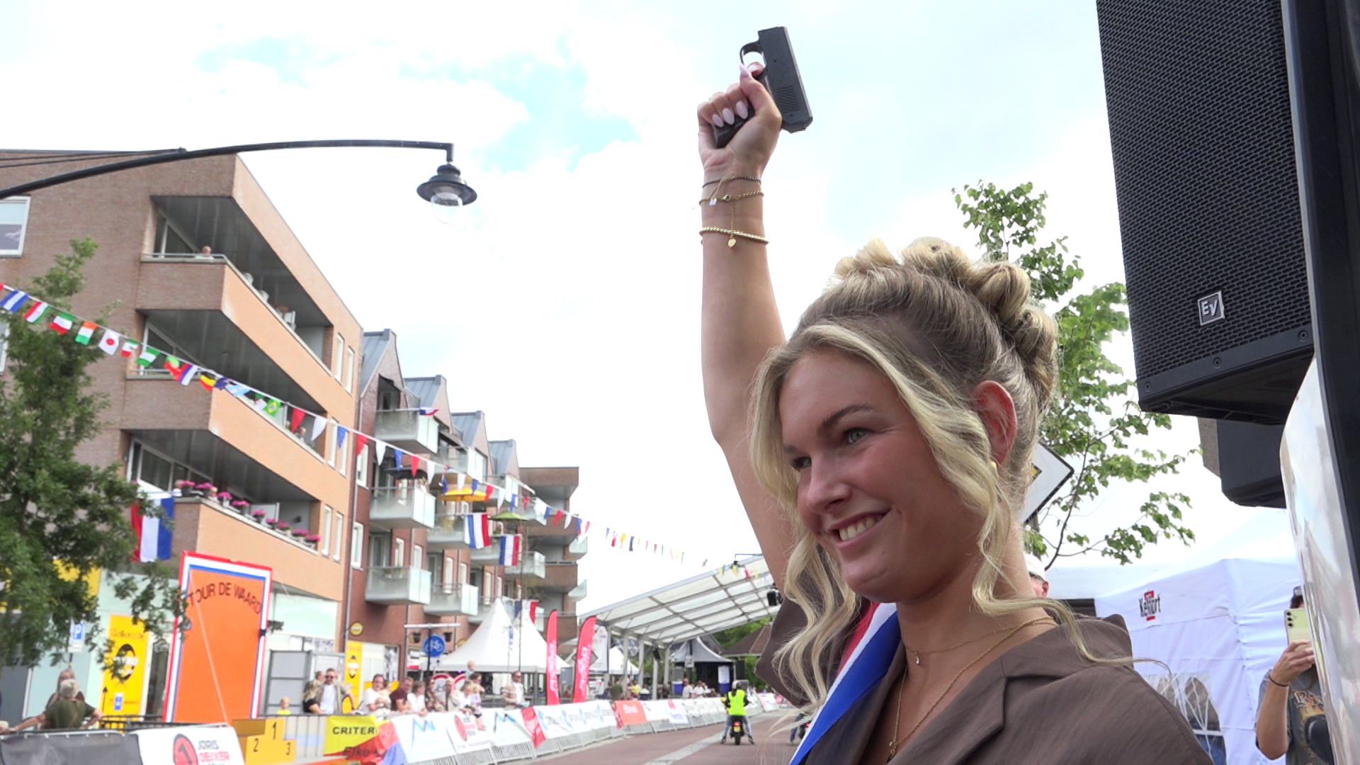 Vrouw met medaille heft een startpistool omhoog bij een wielerwedstrijd op straat met vlaggetjes en publiek
